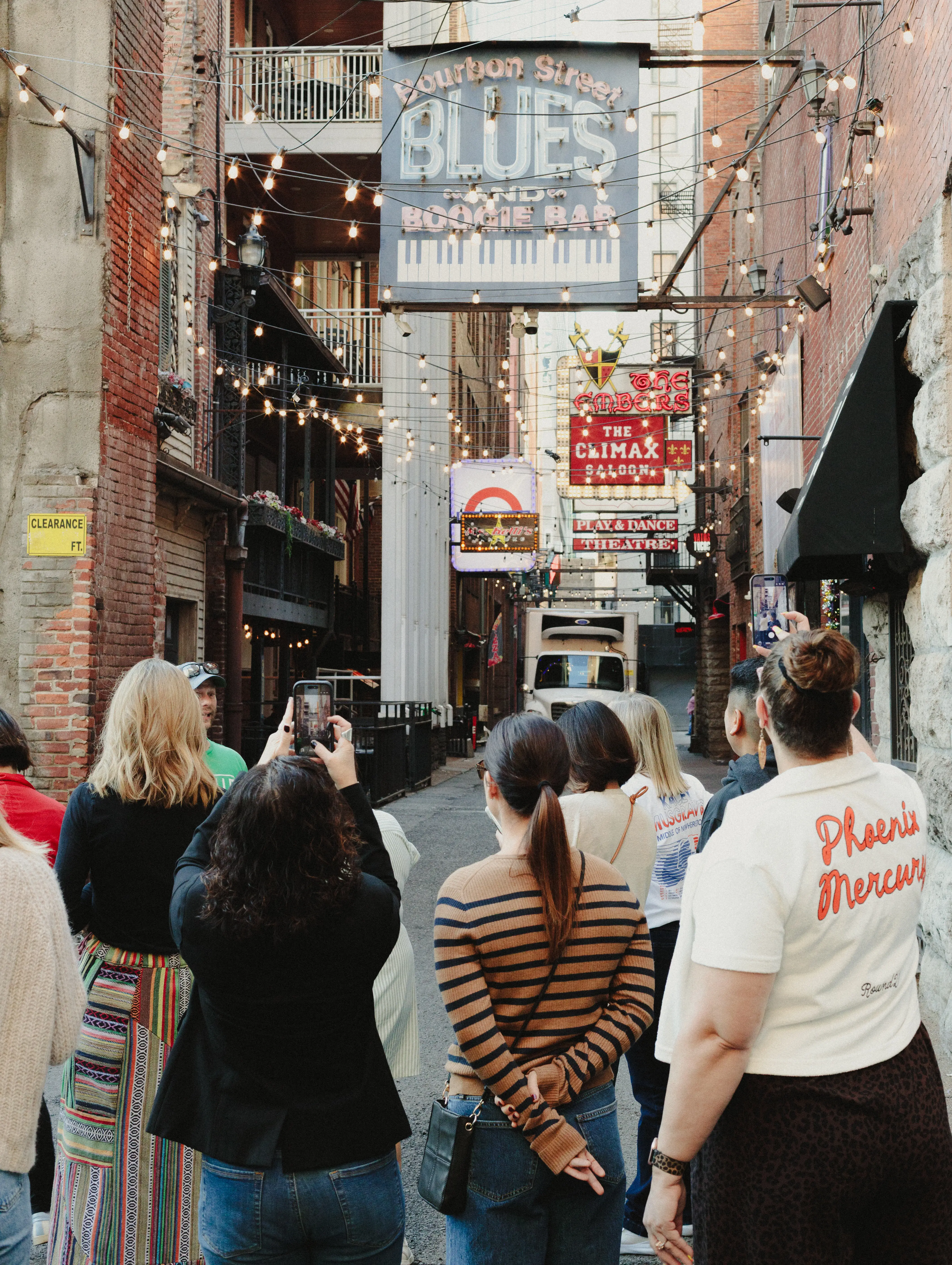 A tour group stands beneath illuminated bar signs and string lights in Printers Alley.