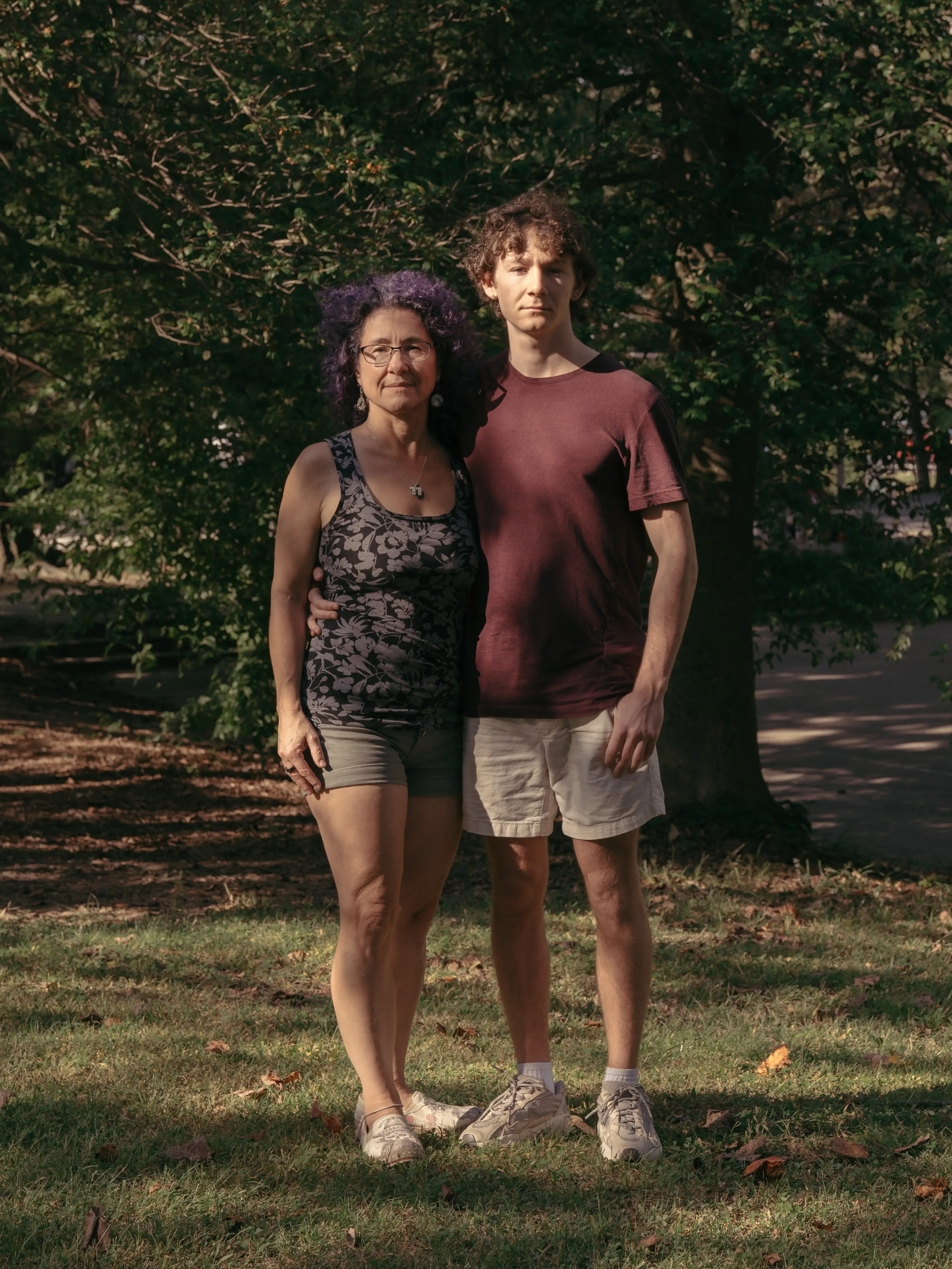 Two people stand with arms around each other on grass in a shaded park.