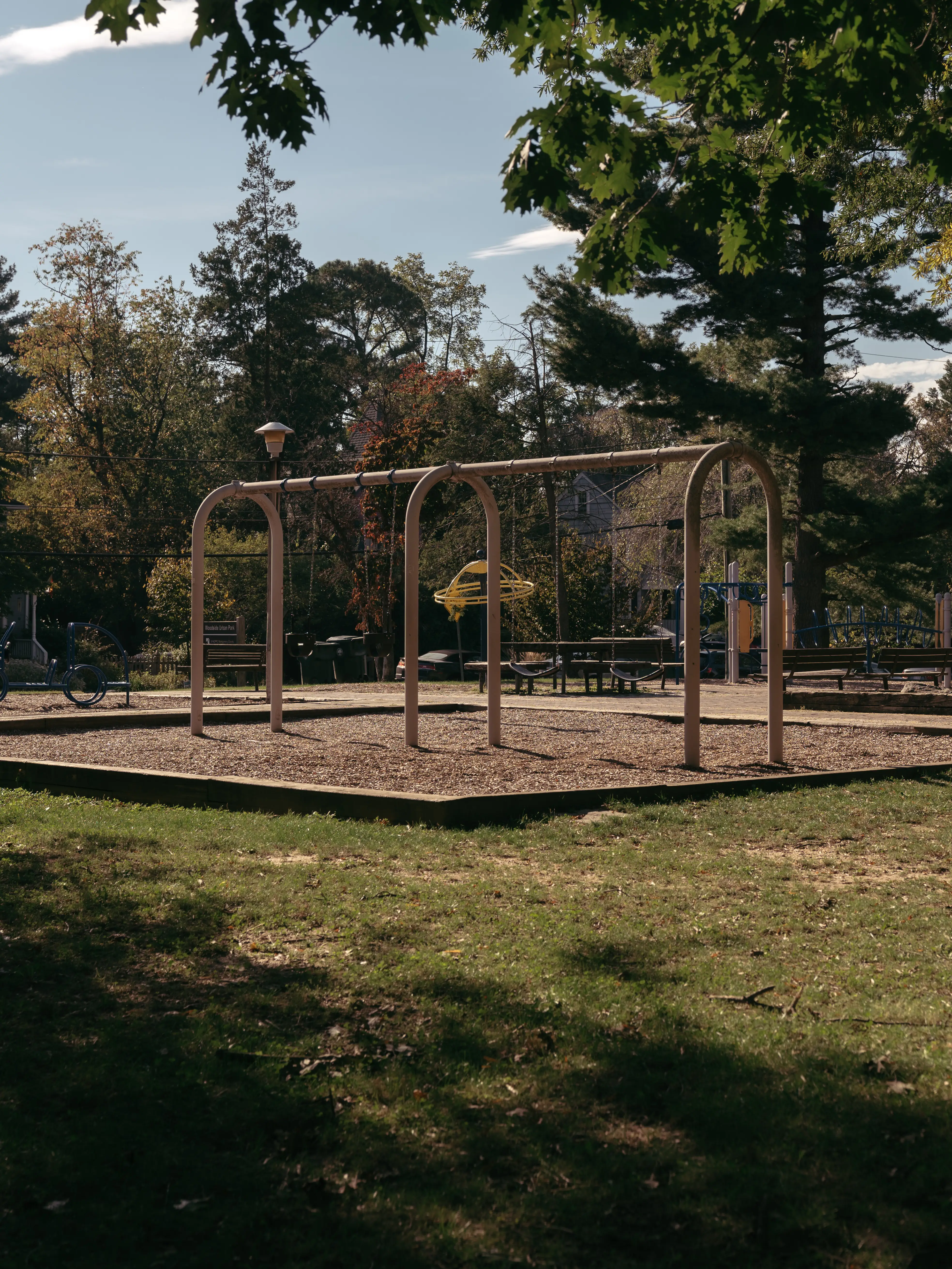 Empty playground swing set and benches sit under leafy trees in a sunny park.