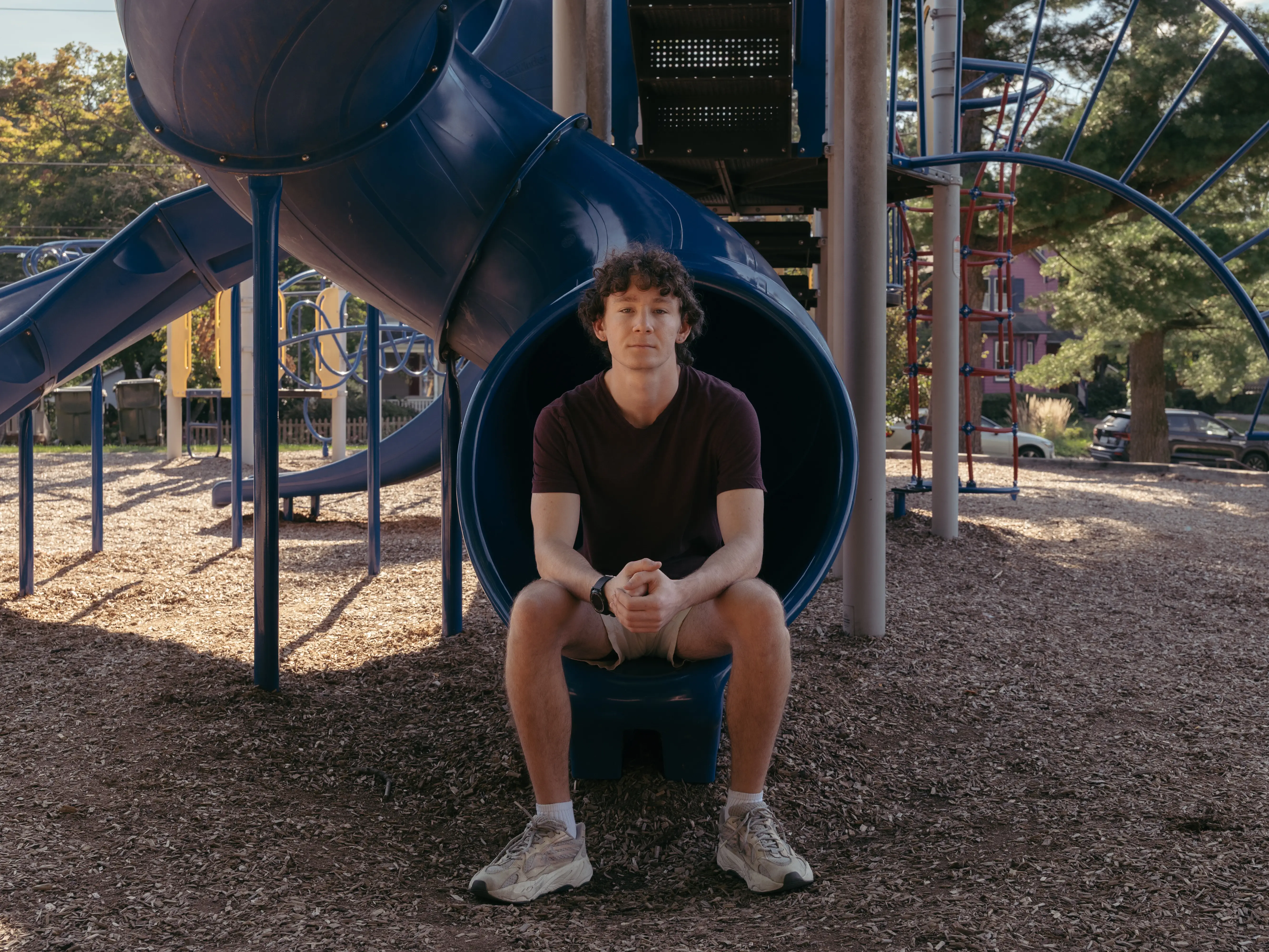 Person seated at the end of a blue tube slide in a playground with climbing structures and trees.