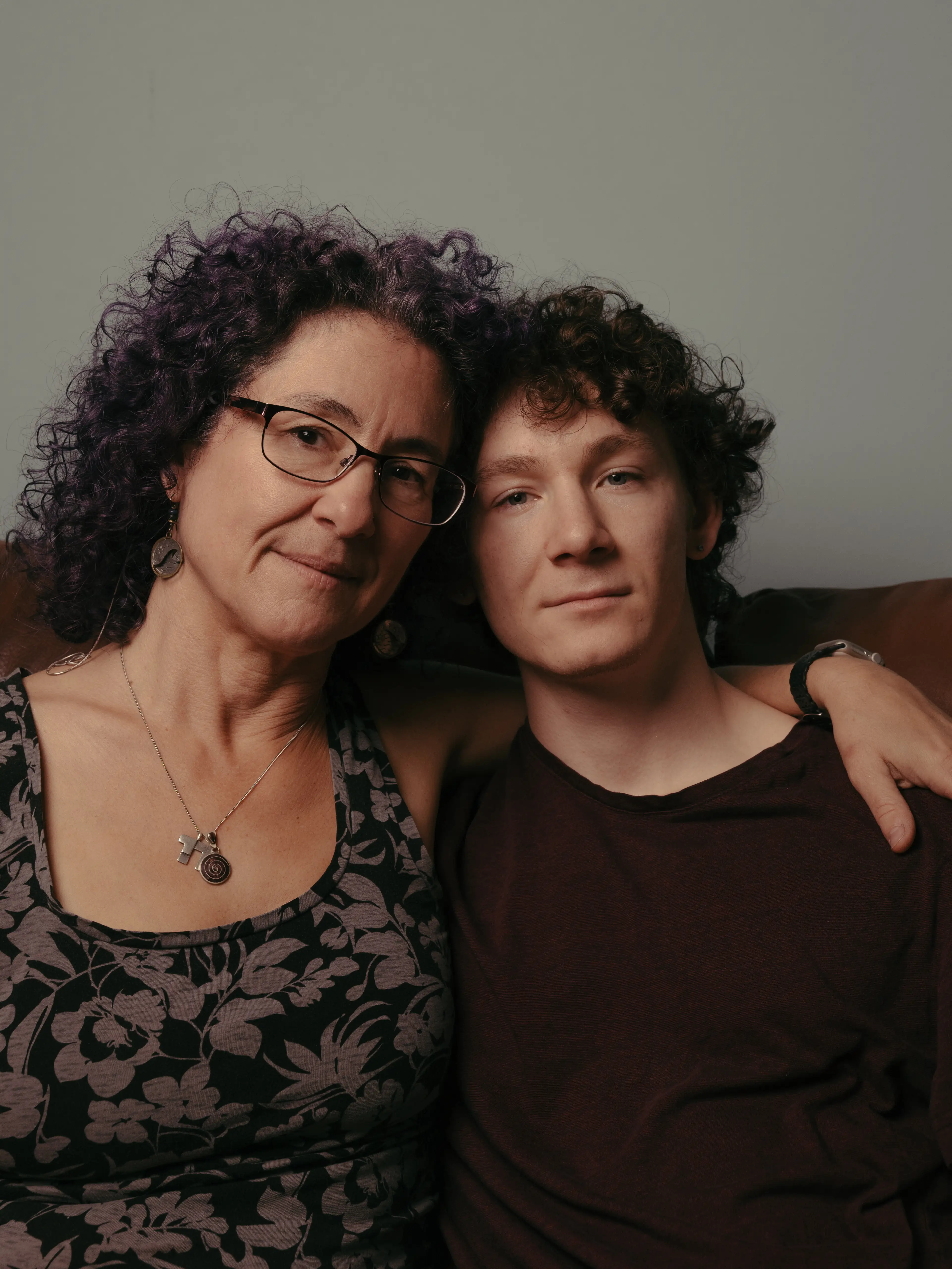 Two people with curly hair sit closely together on a couch, one wearing glasses and patterned clothing.
