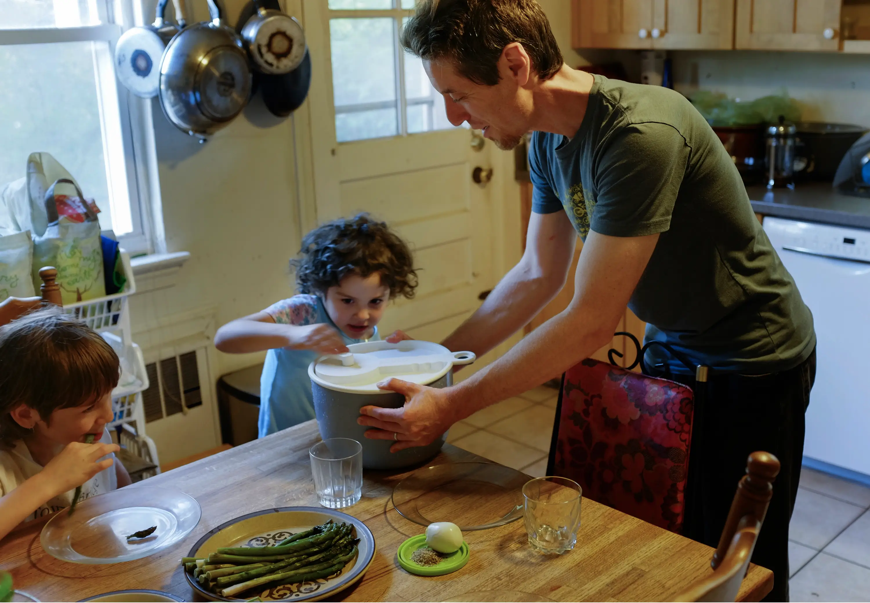 One person helps two seated people dish food at a kitchen table with asparagus and glasses.