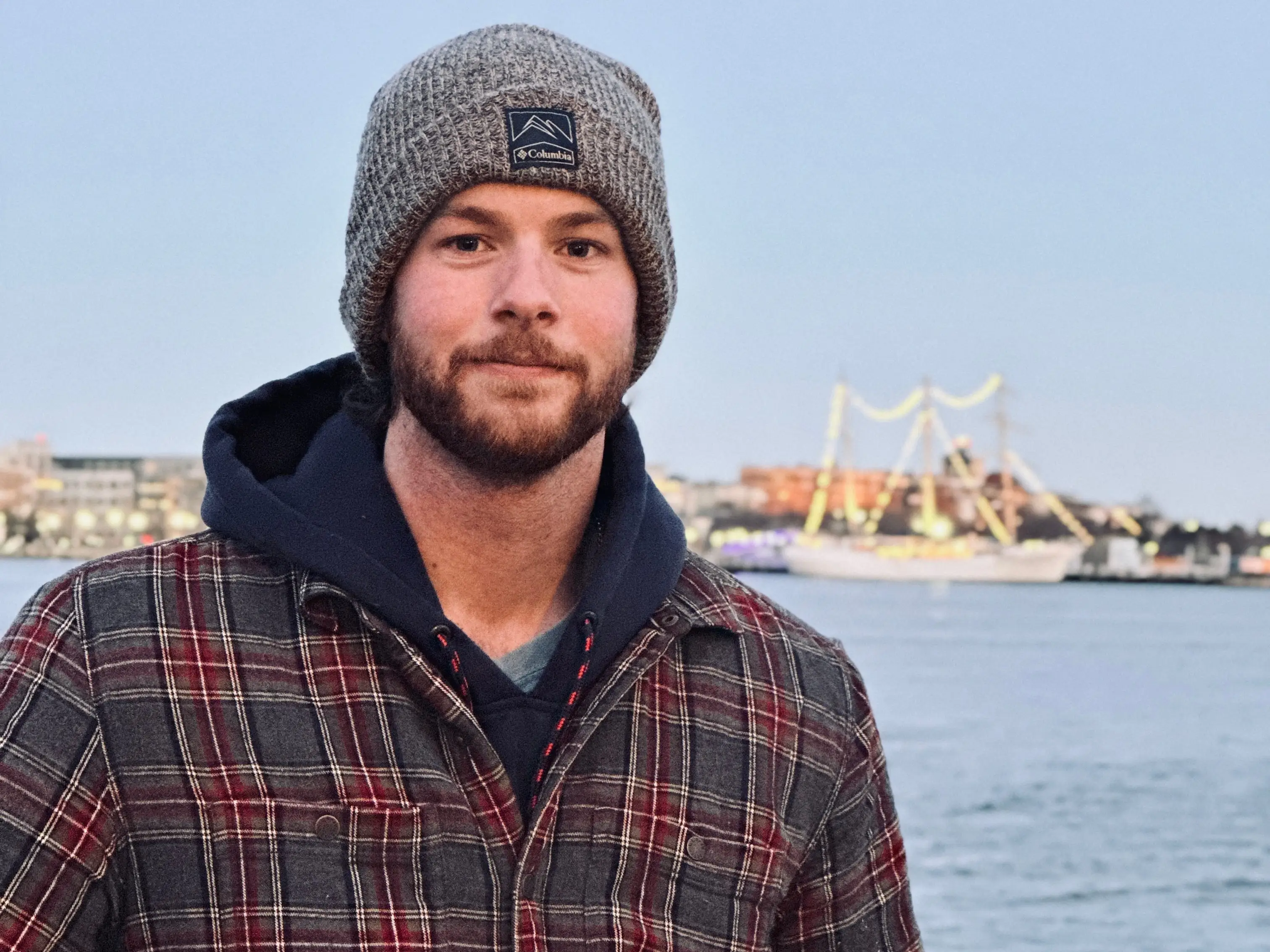 Joseph LeGare standing outdoors by a rail with water in the background, wearing a plaid jacket and a skull cap.