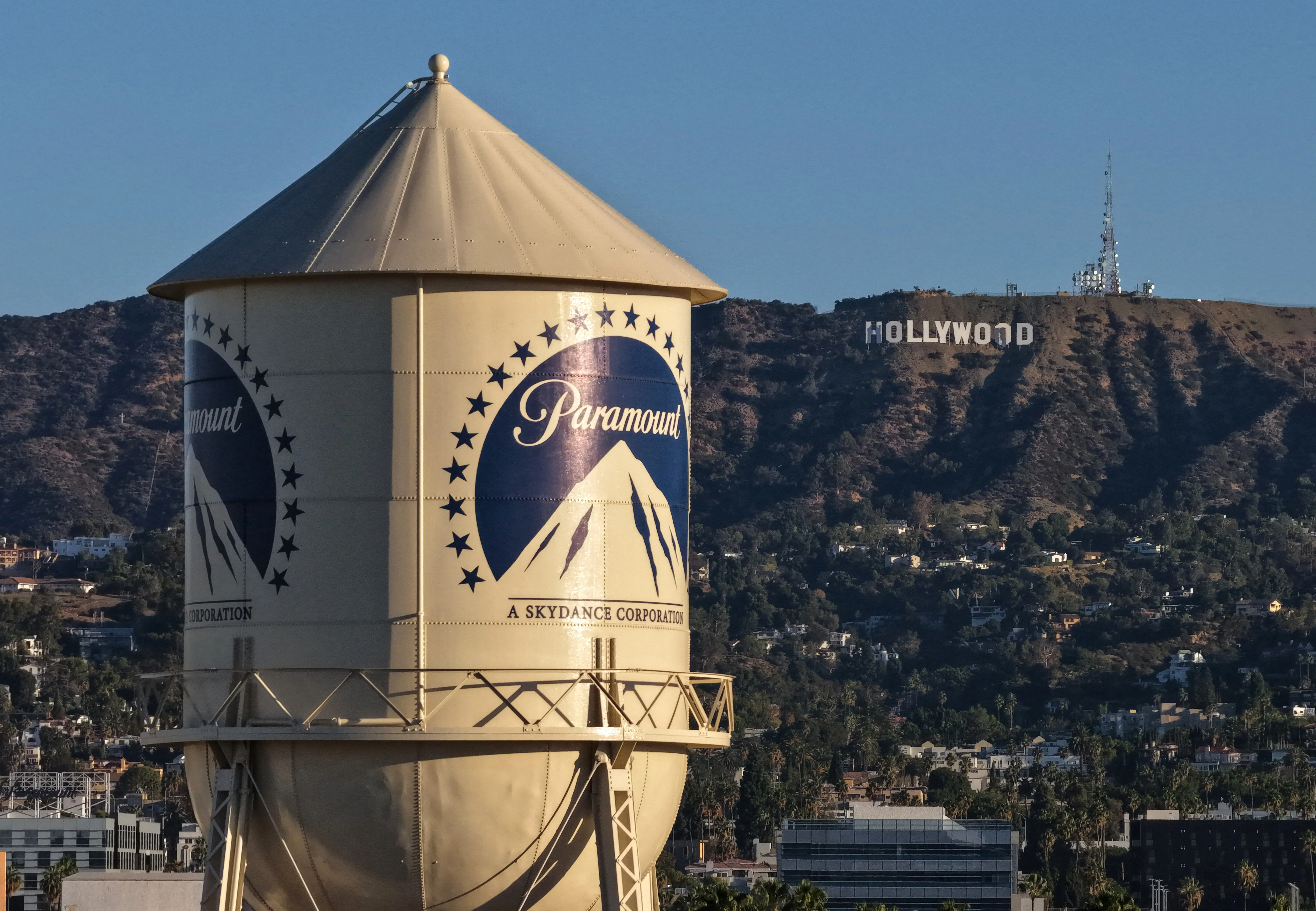 Paramount water tower stands in front of the Hollywood Sign on a hillside under a clear blue sky.