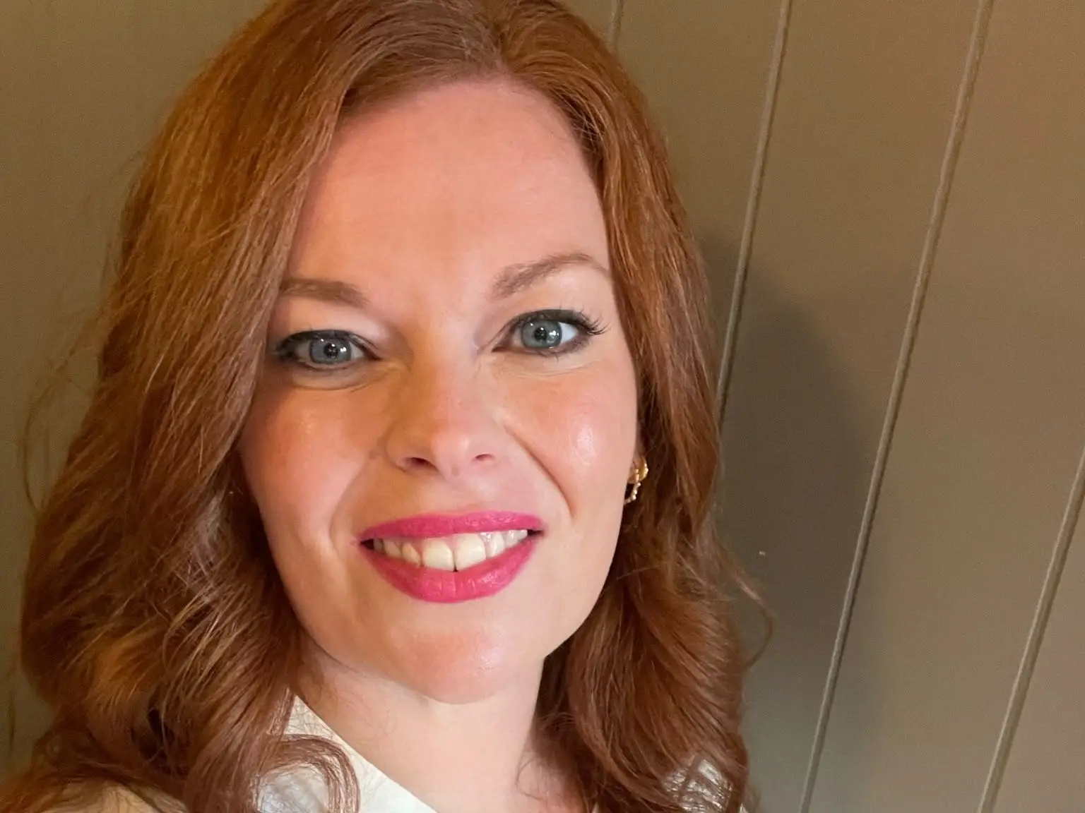 A smiling red-haired person in a white button-up shirt poses for a close-up selfie indoors.
