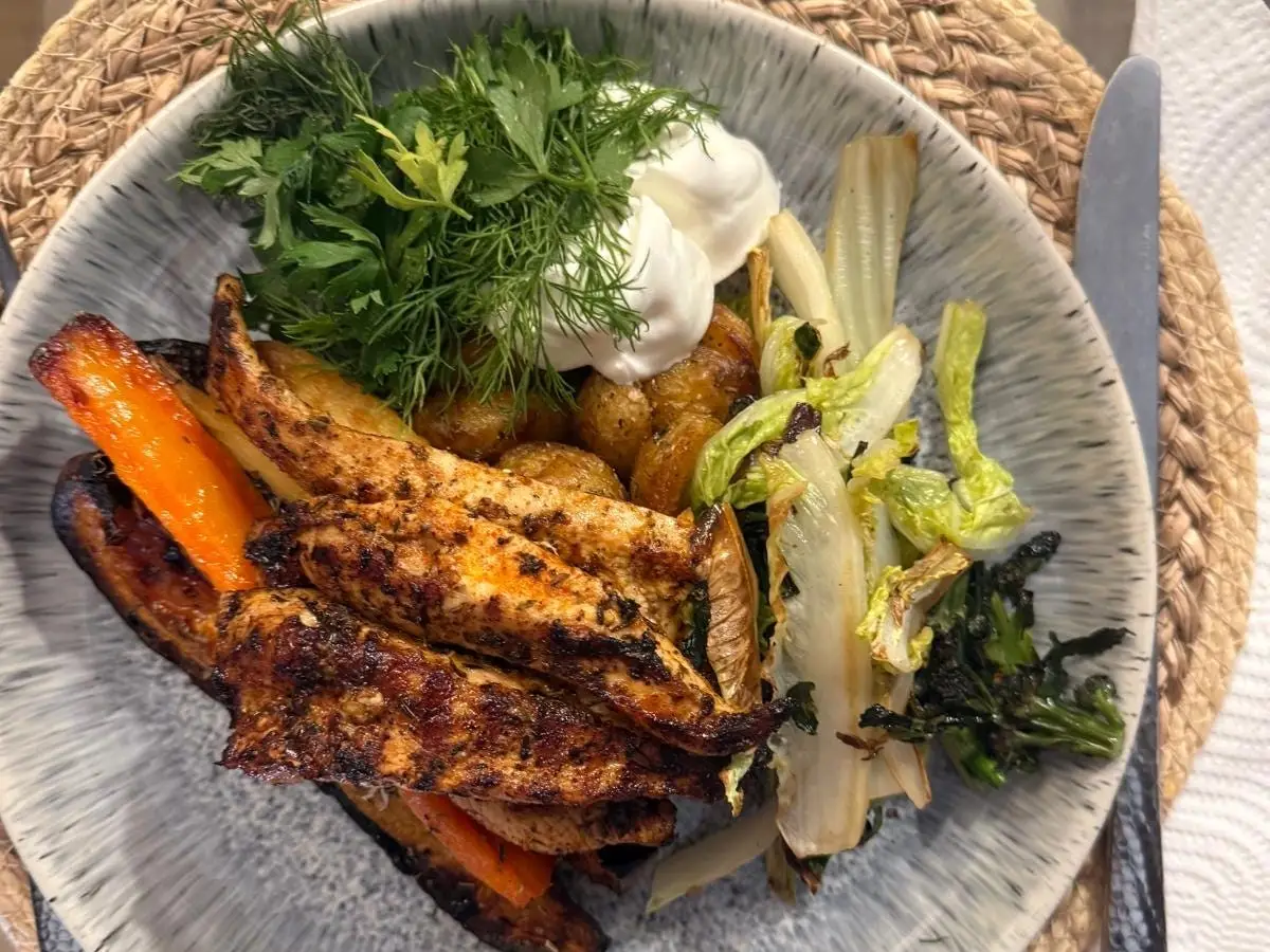 Bowl of grilled meat, roasted vegetables, potatoes, herbs, and white sauce on a woven placemat.
