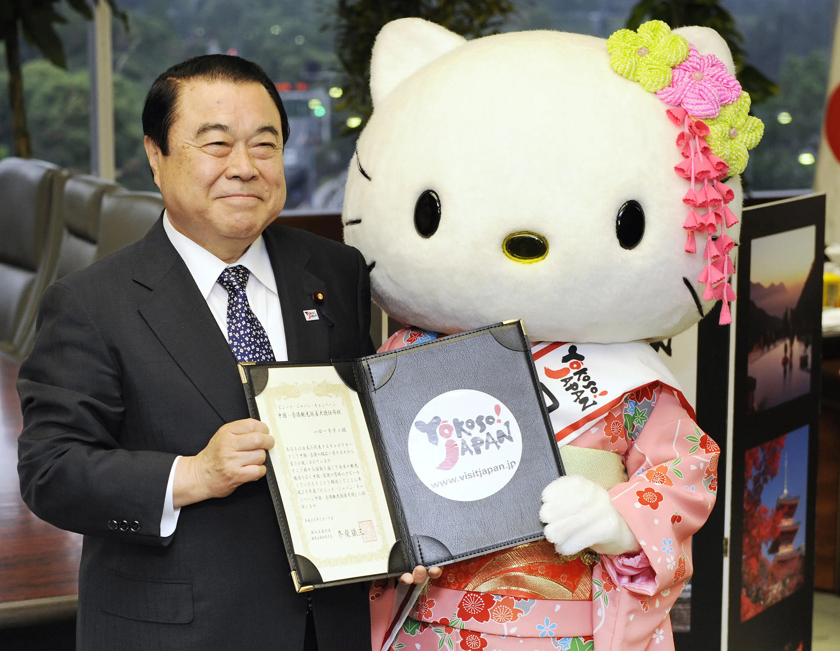 A suited person and Hello Kitty mascot hold an open certificate and Yokoso! Japan folder indoors.