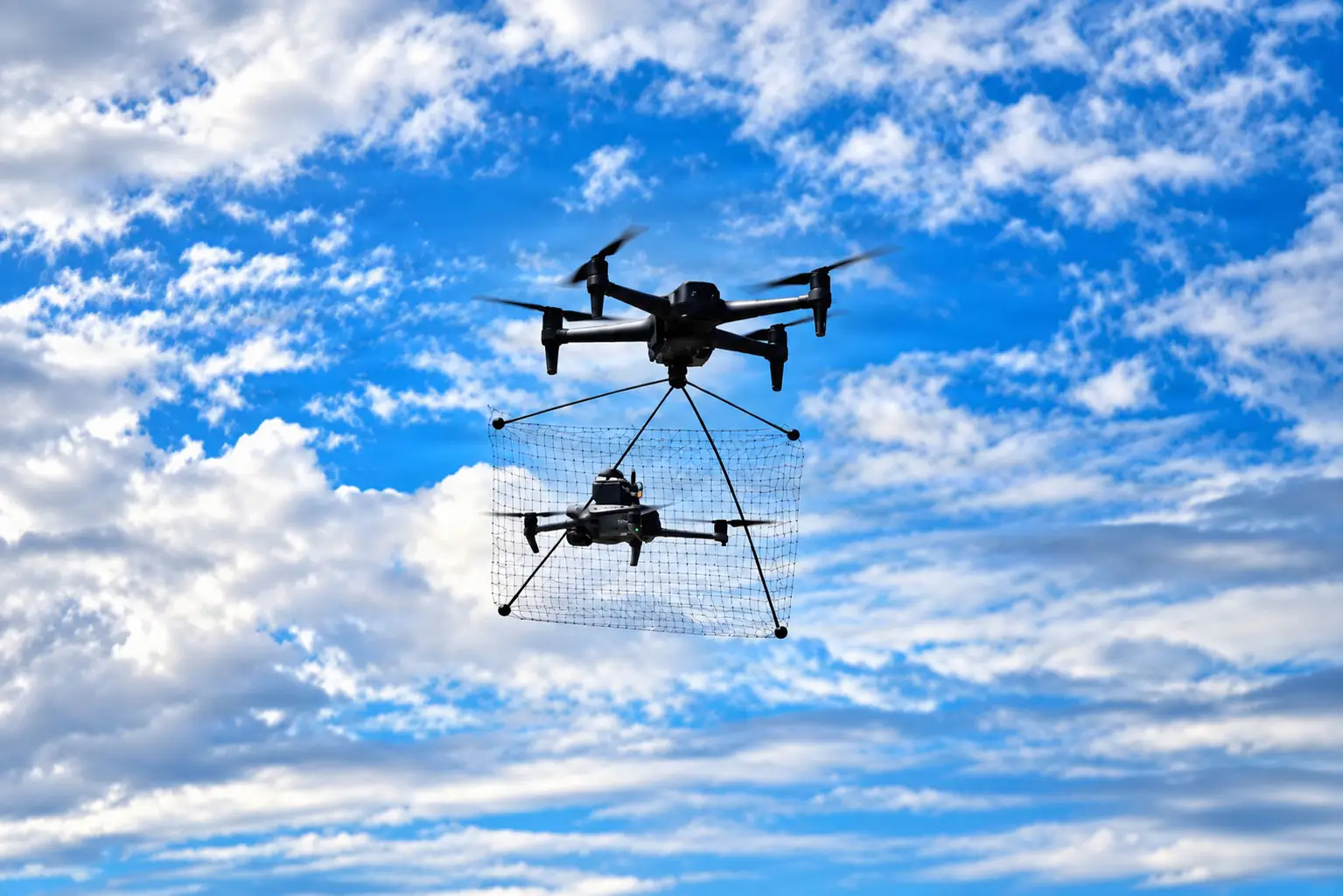 A quadcopter is seen using its net to catch a drone in a bright blue and cloudy sky.
