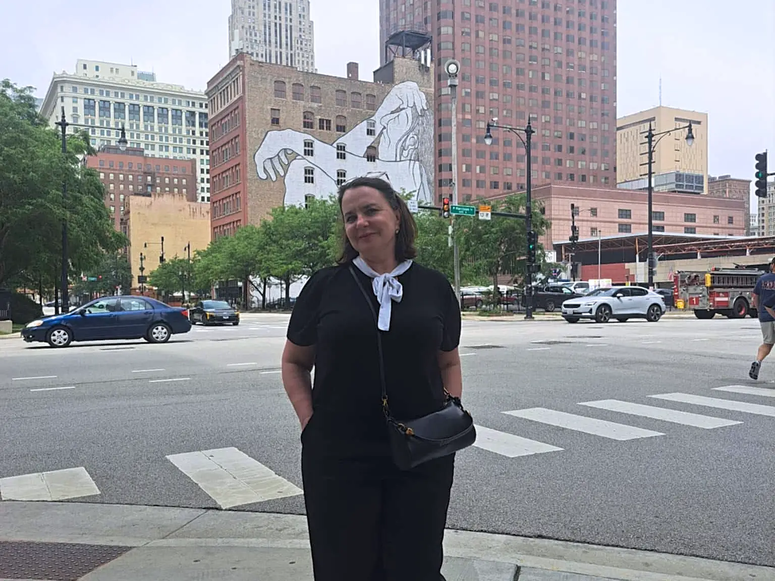 A person in black clothing stands on a downtown sidewalk near a crosswalk, traffic, skyscrapers, and a large mural.
