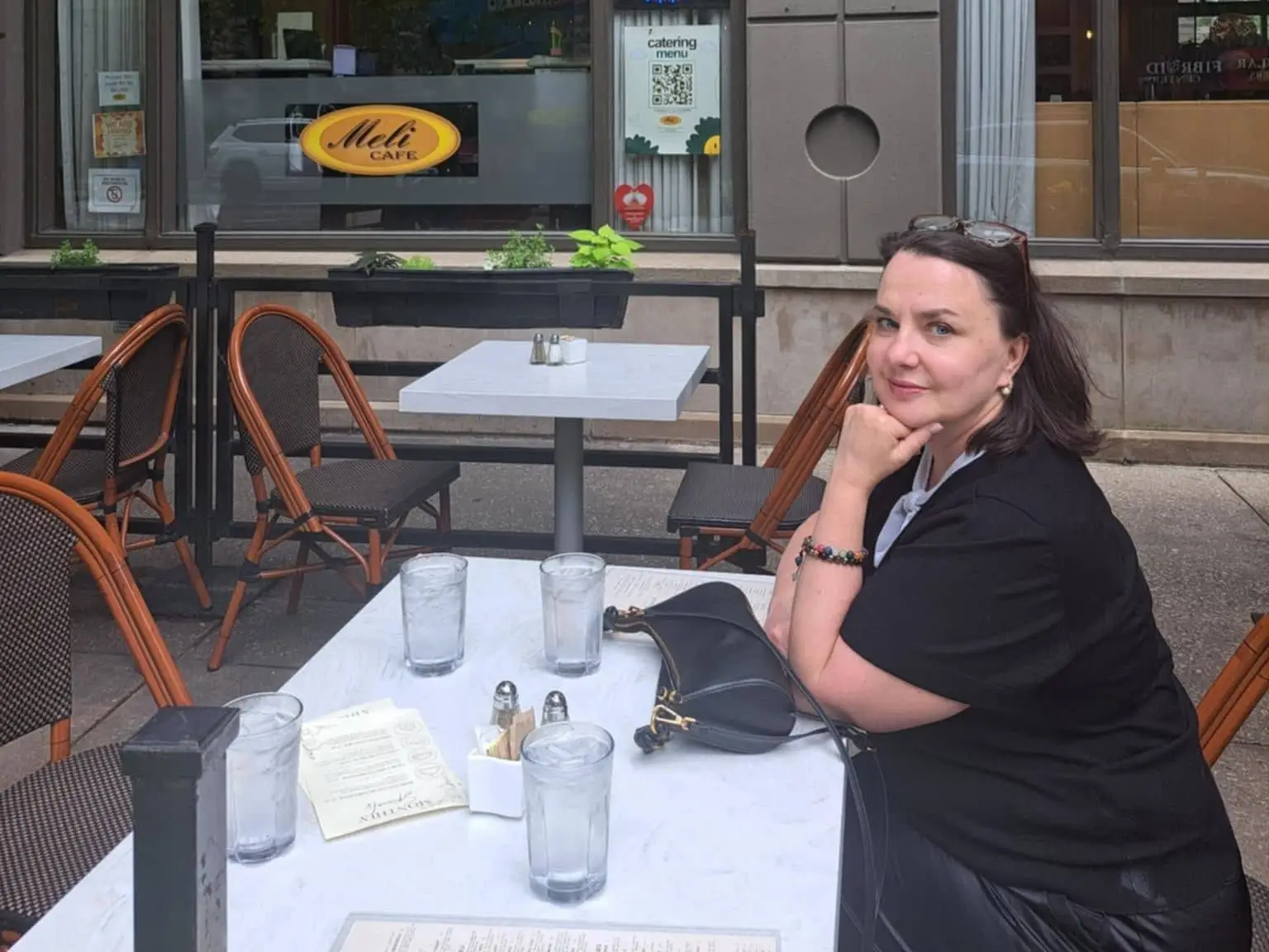 Person seated at an outdoor café table in front of Meli CAFE windows with an OPEN sign.