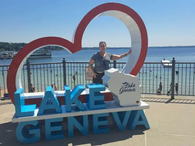 Person poses beside a large Lake Geneva waterfront sign with a lake, beach, and pier in the background.