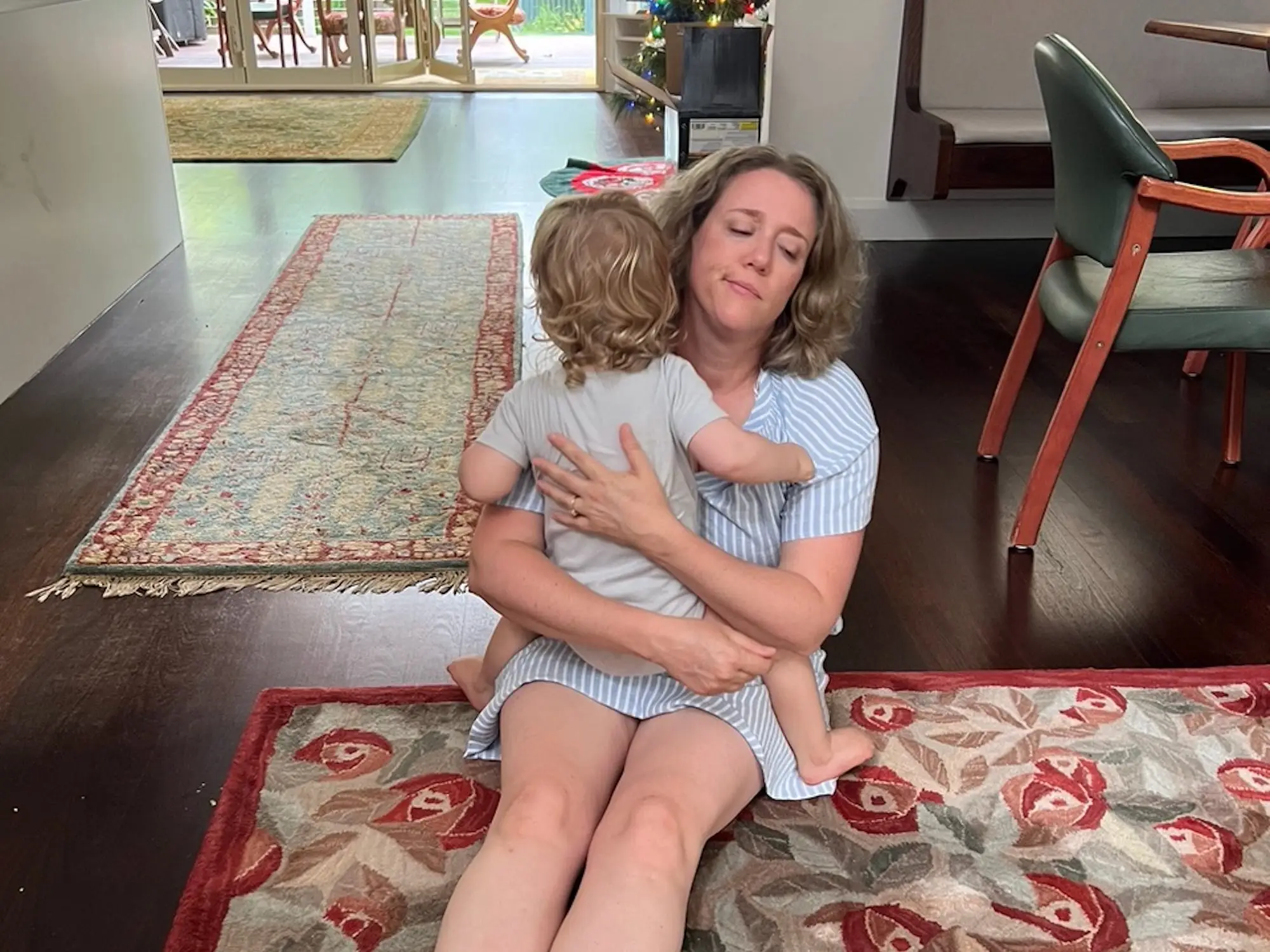 Adult seated on a patterned rug hugs a small child in a home living room with a decorated tree.