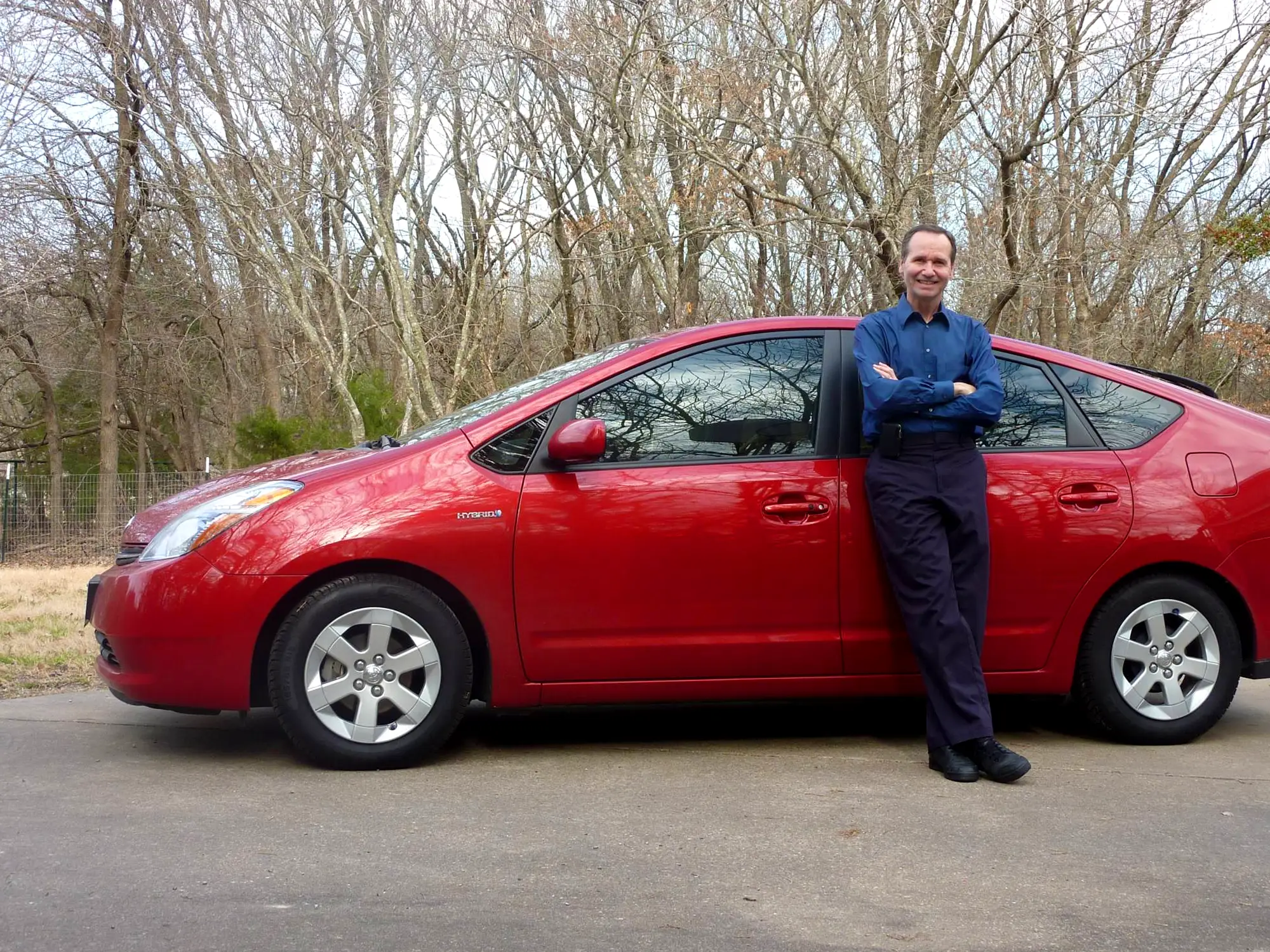 Person standing with arms crossed beside a red Toyota Prius on a paved road near leafless trees.