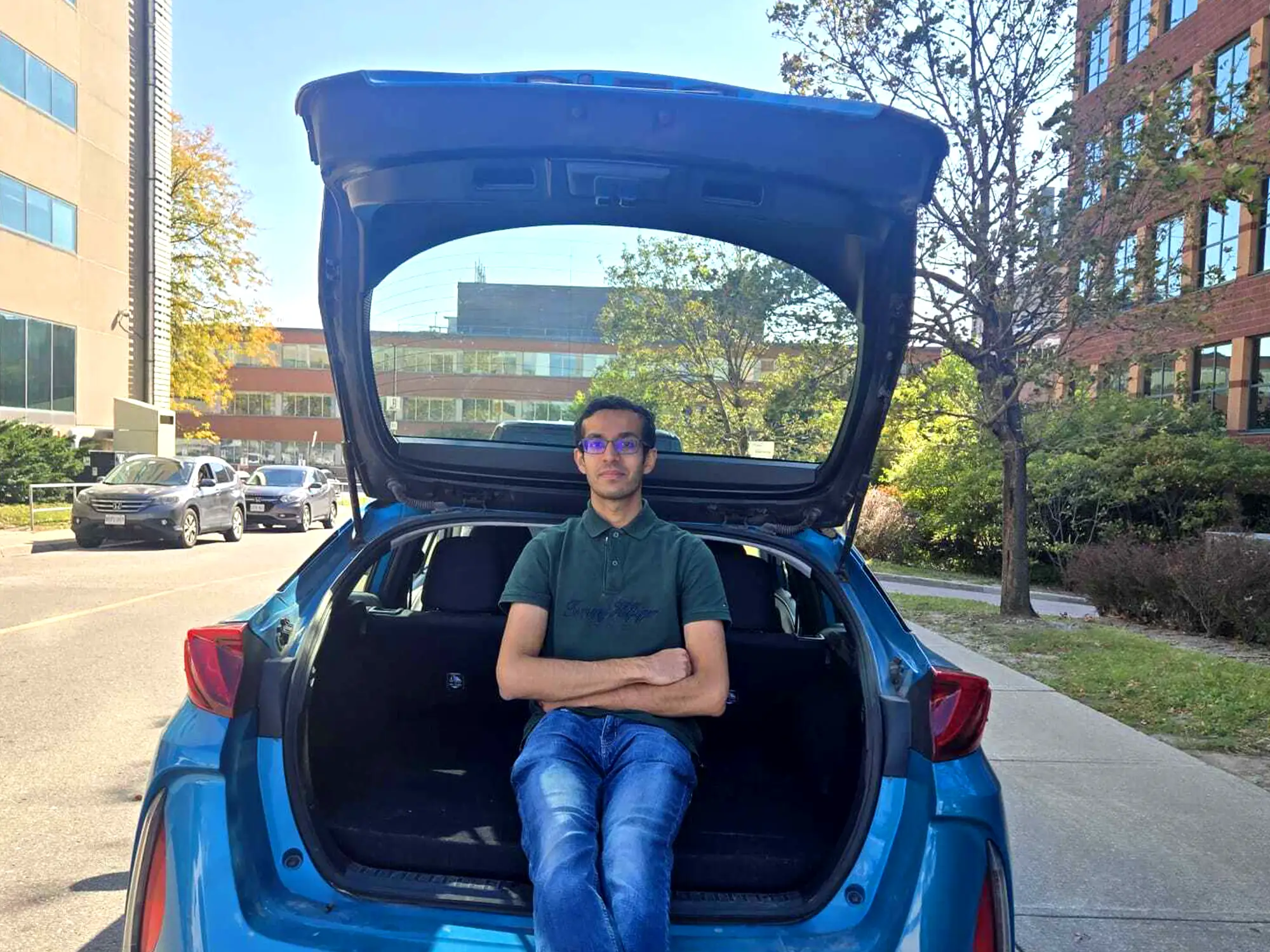 A person wearing sunglasses sits in the open trunk of a blue hatchback parked on an urban street.