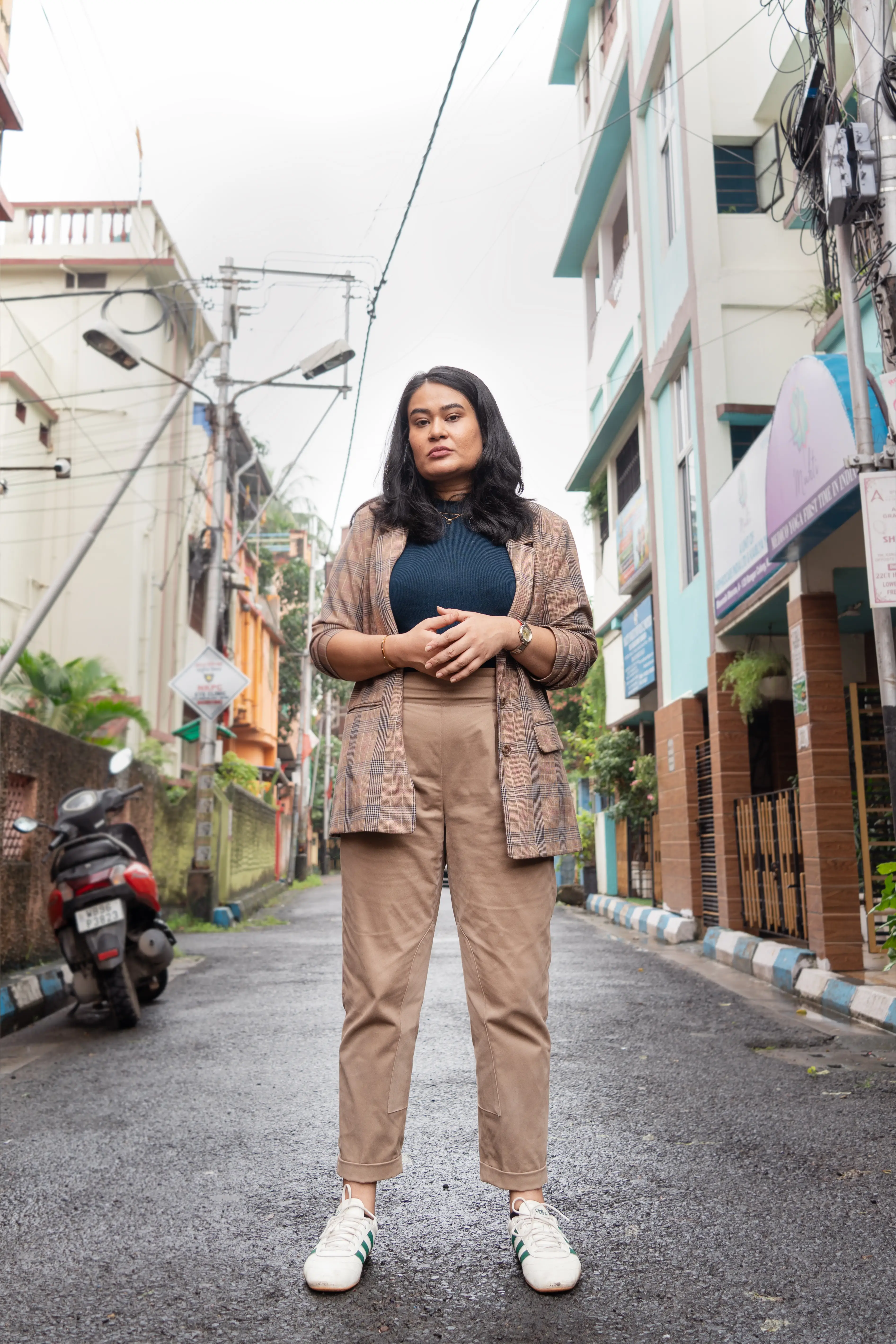 Person stands in a narrow urban street lined with buildings, parked scooter, overhead wires, and streetlights.