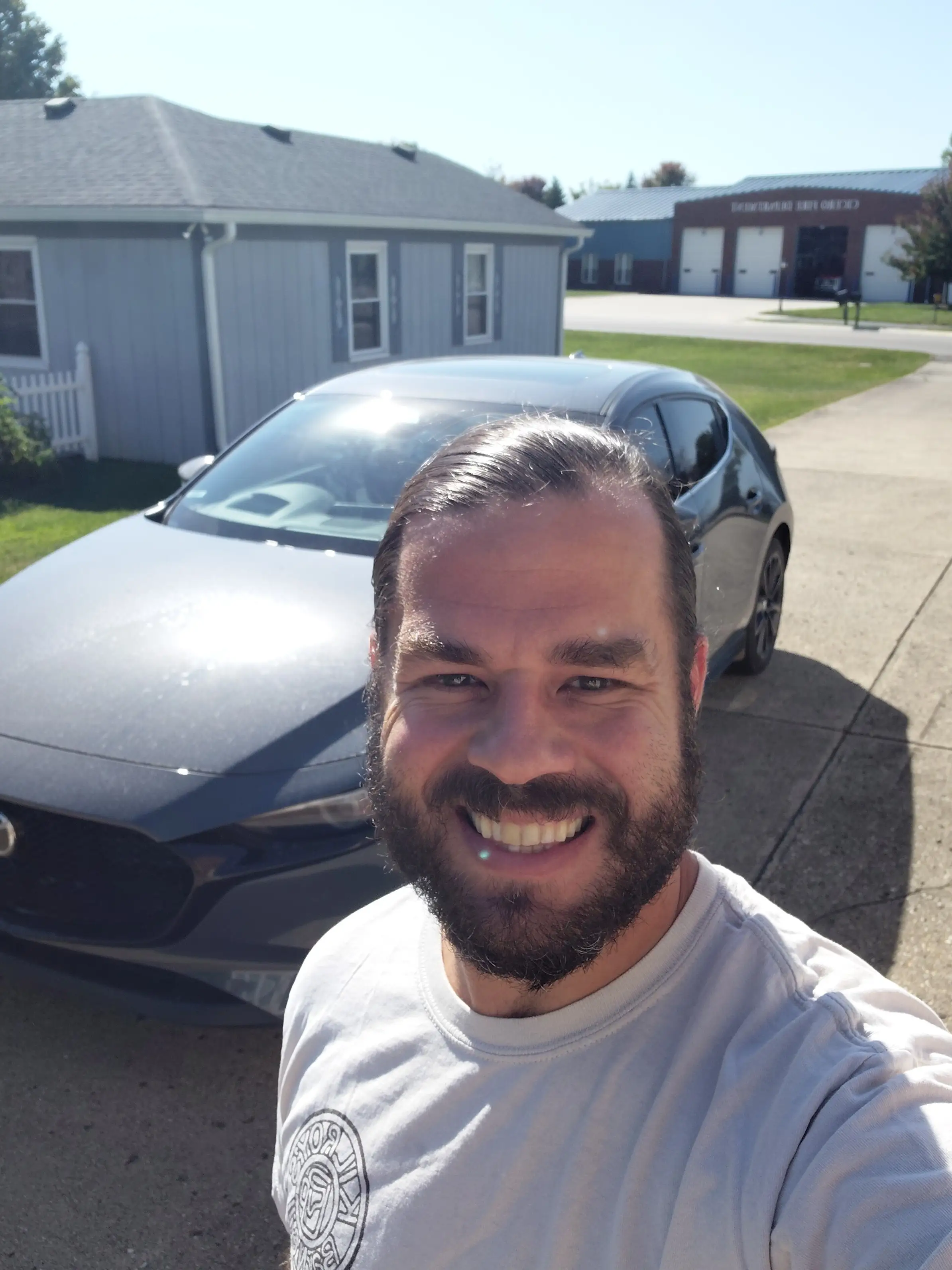 Smiling person takes a selfie in a driveway with a gray Mazda car and township road center building behind.
