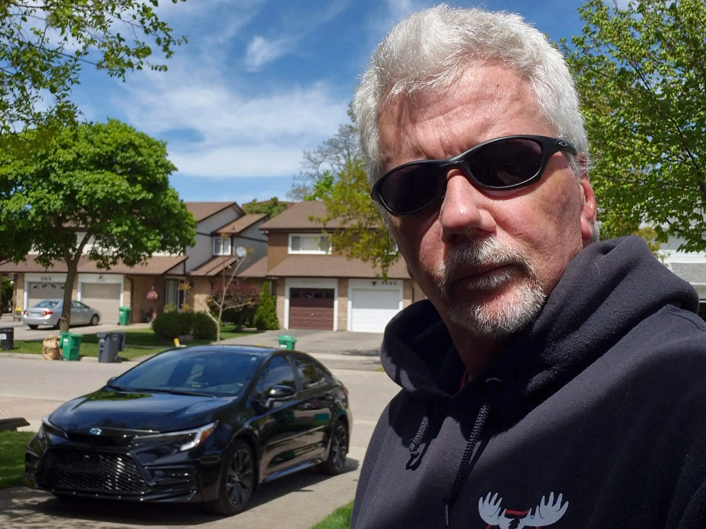 Person in sunglasses and a dark hoodie stands on a suburban street beside a black Toyota sedan.