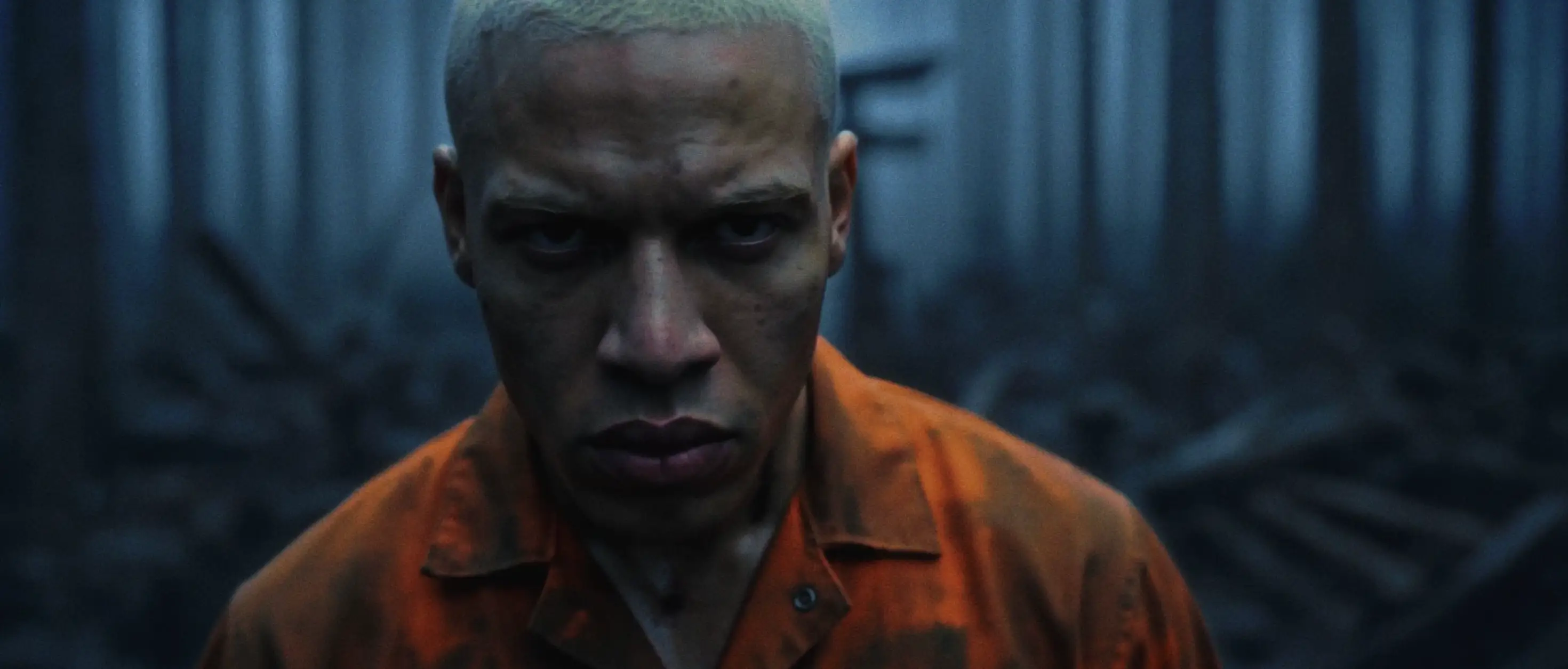 Close-up of a person in an orange shirt staring intensely in a dark industrial corridor.