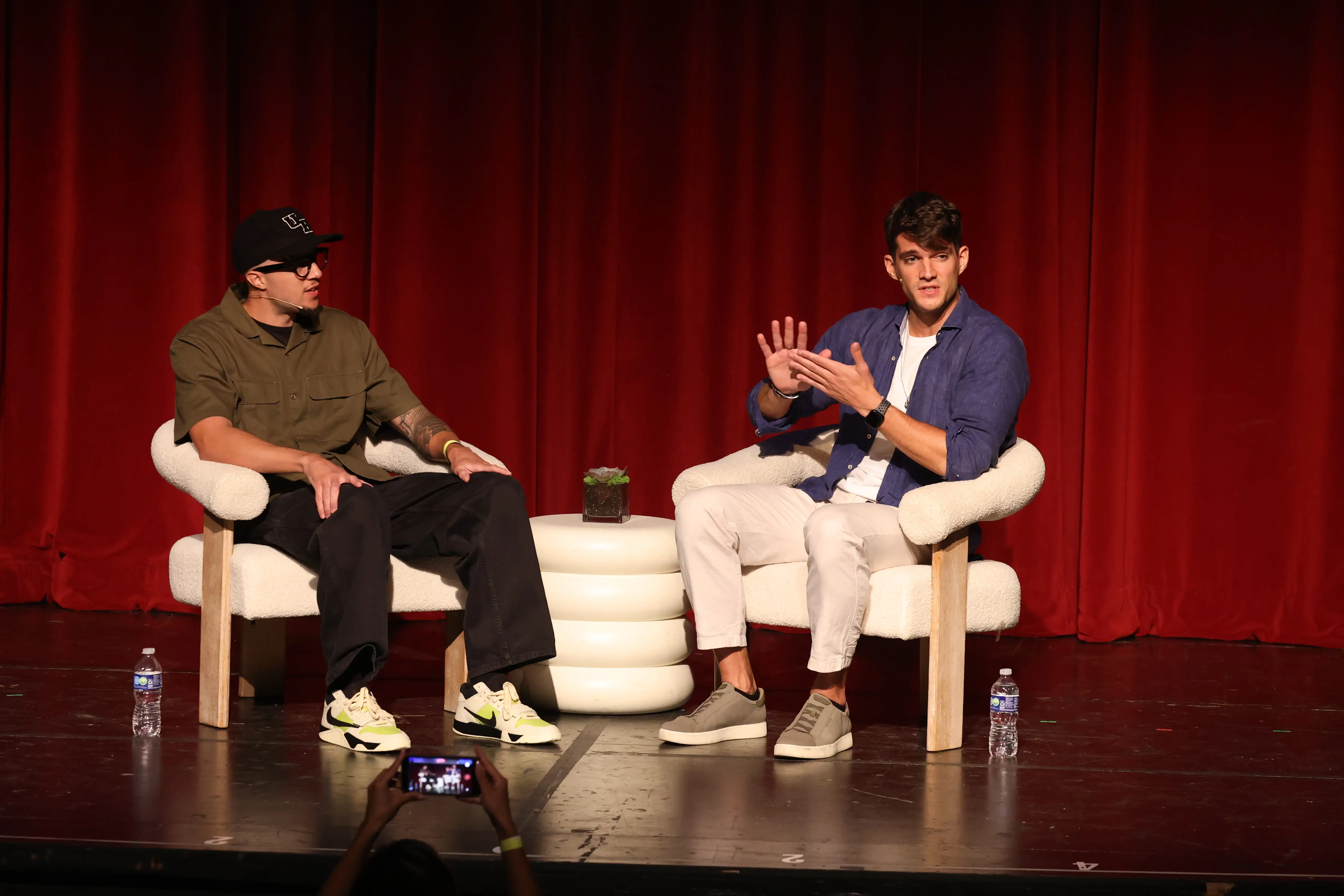 Two speakers sit in armchairs on a stage before a red curtain during a live audience event.
