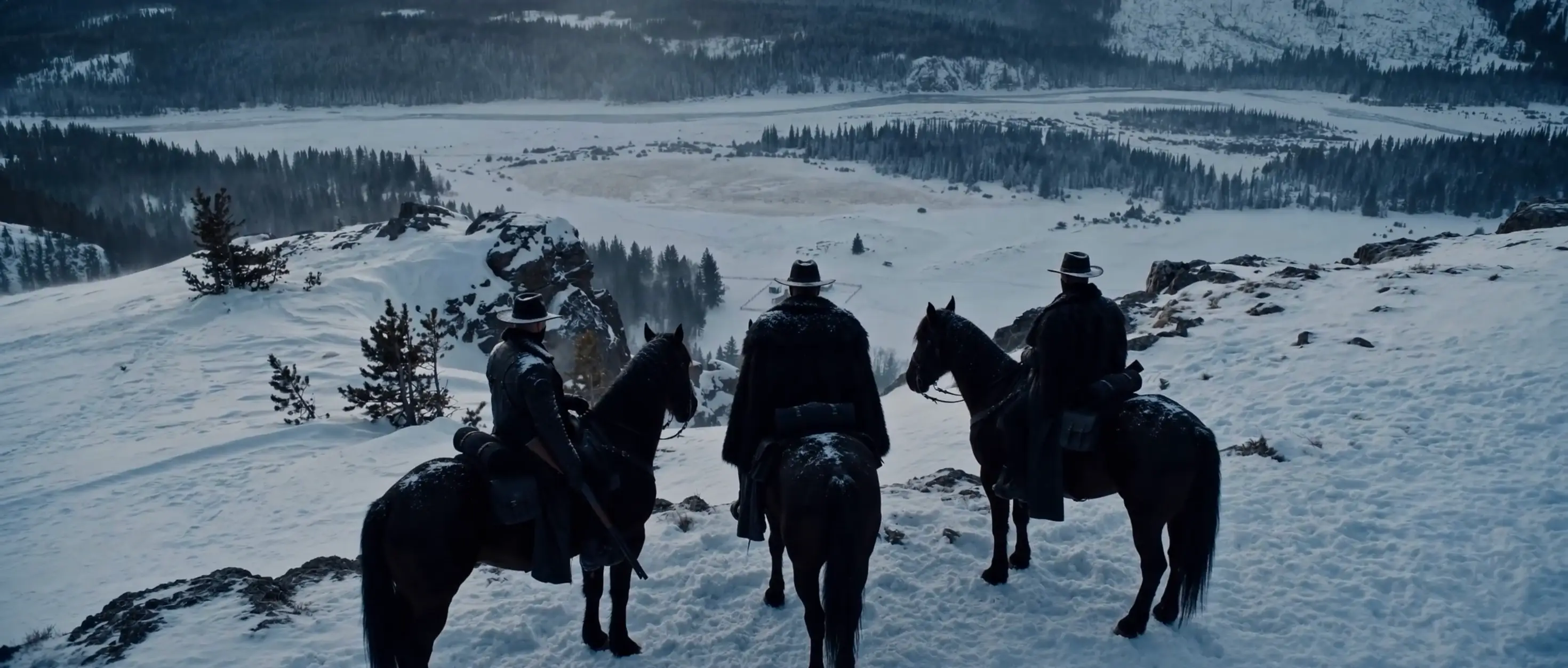 Three horseback riders overlook a snowy mountain valley from a high ridge.