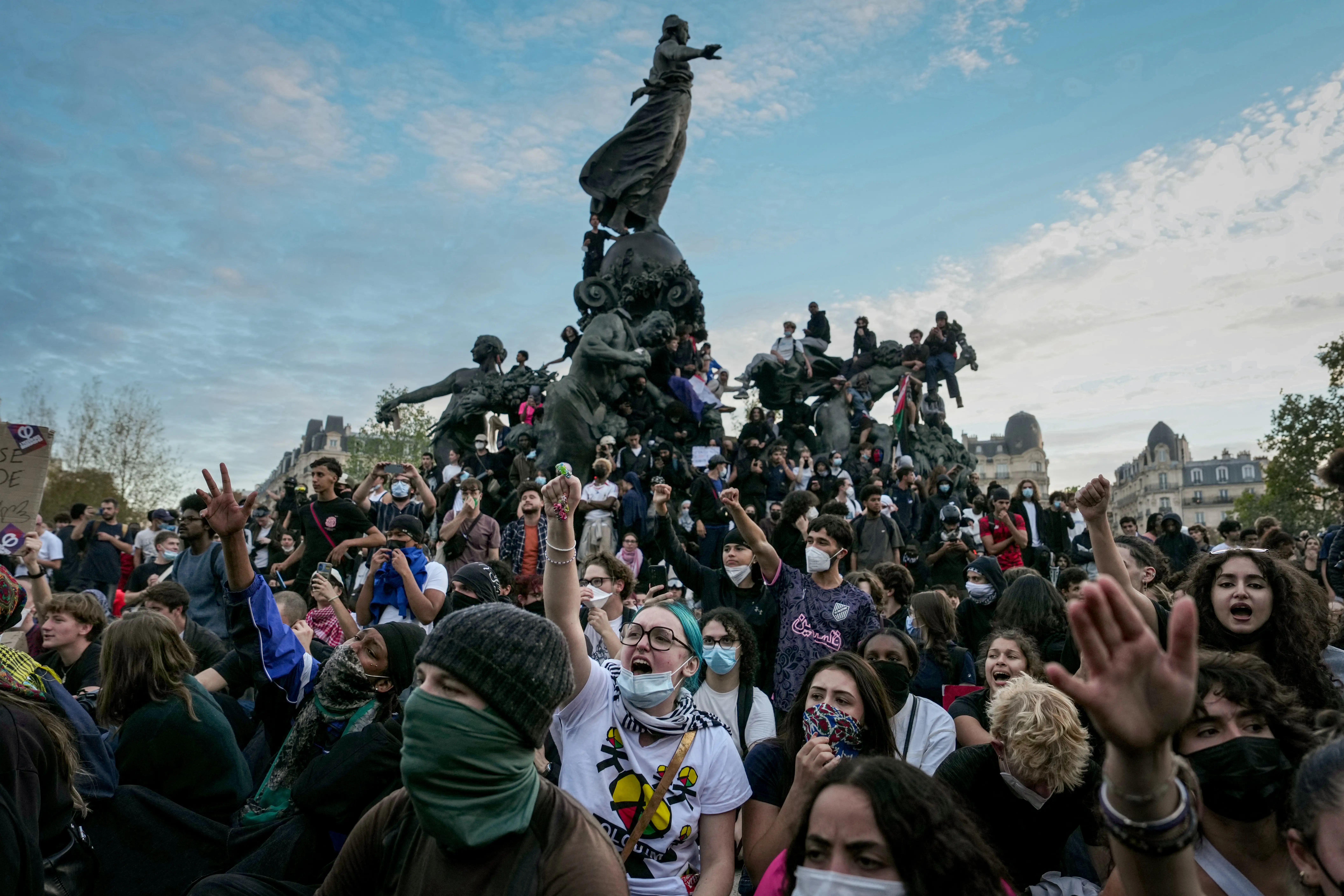 Crowd of masked demonstrators raises hands around the Monument à la République in a city square.