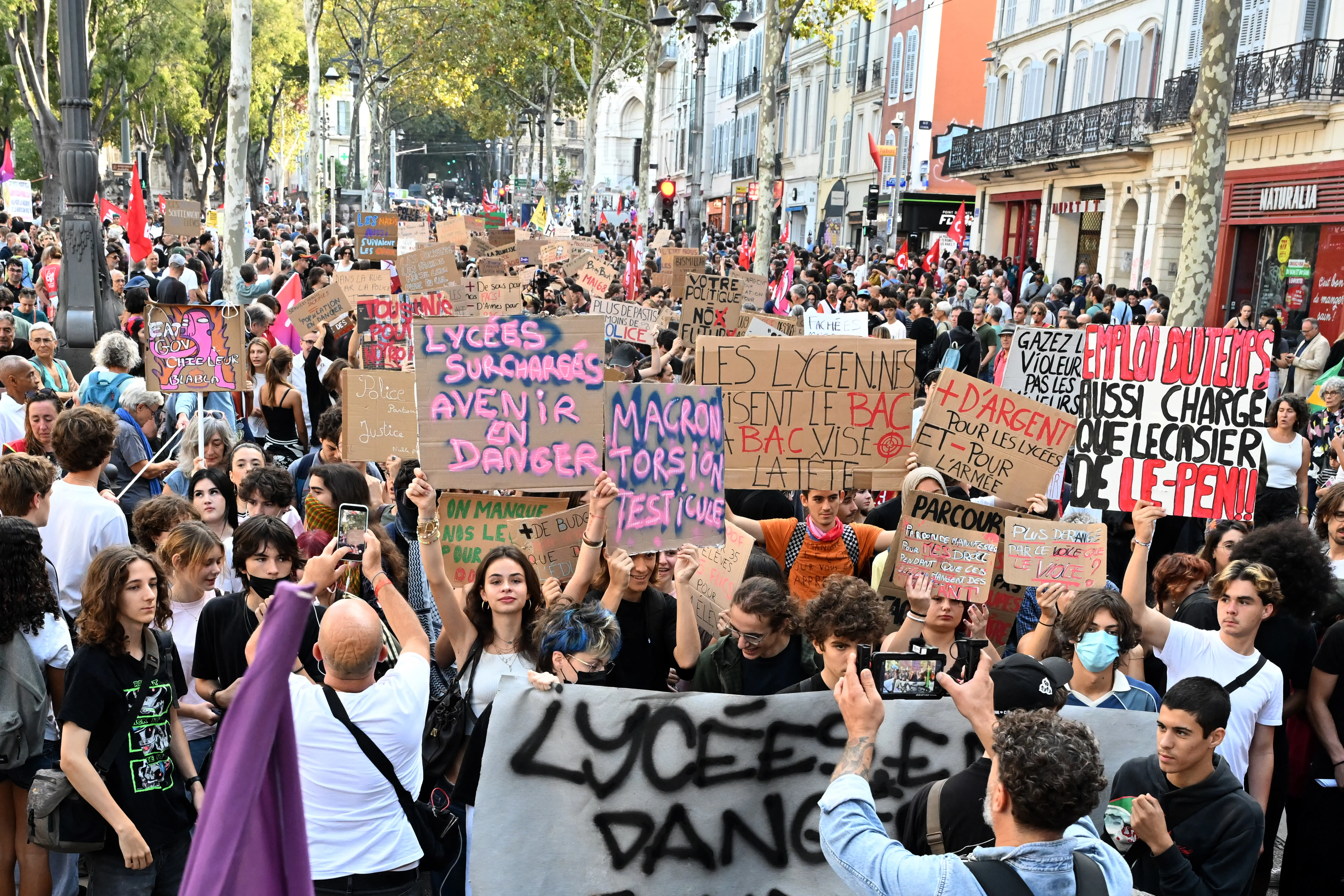 Crowd of demonstrators fills a tree-lined city street holding French protest signs about overcrowded high schools.