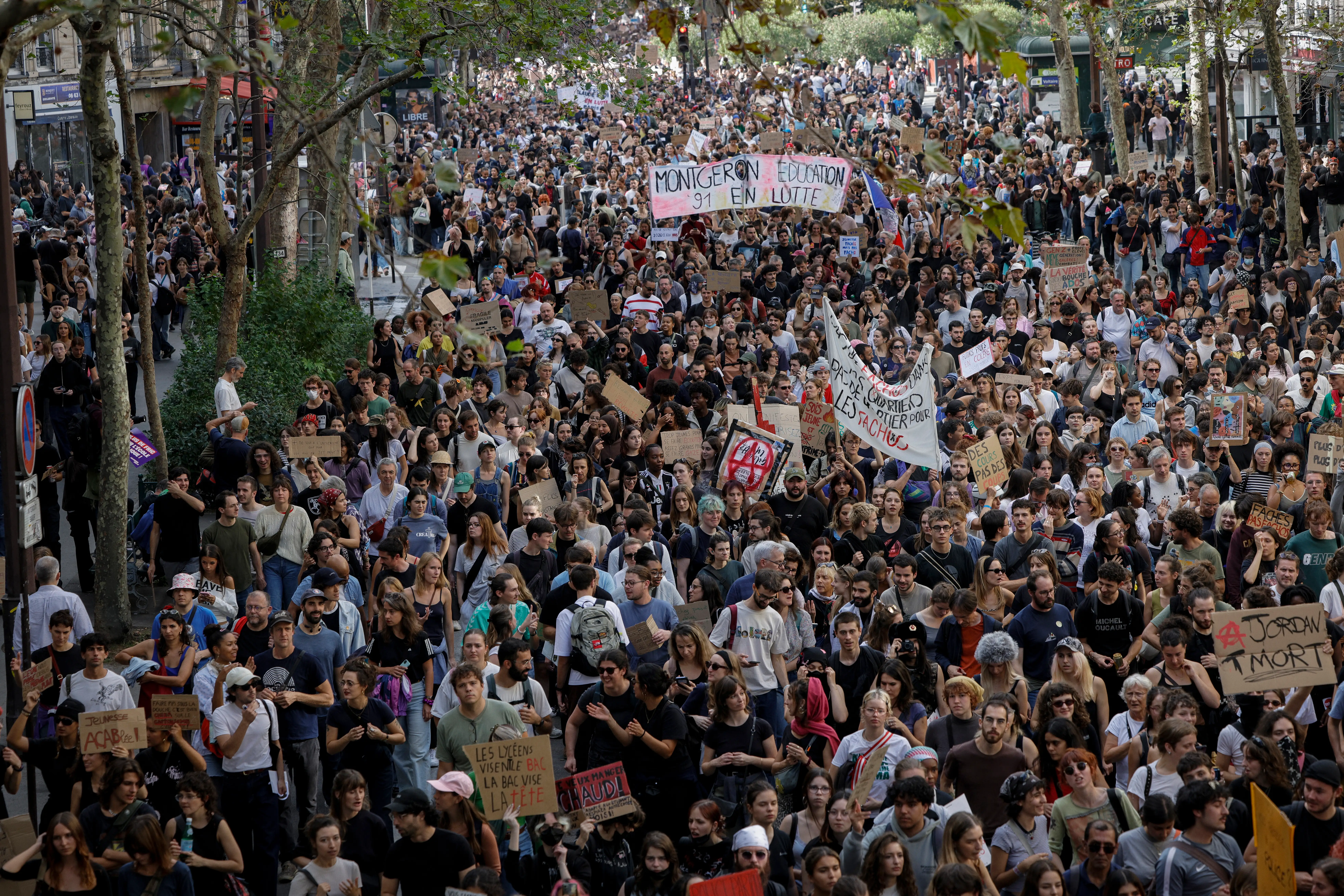 Crowd fills a tree-lined city street during a public march with banners and protest signs.