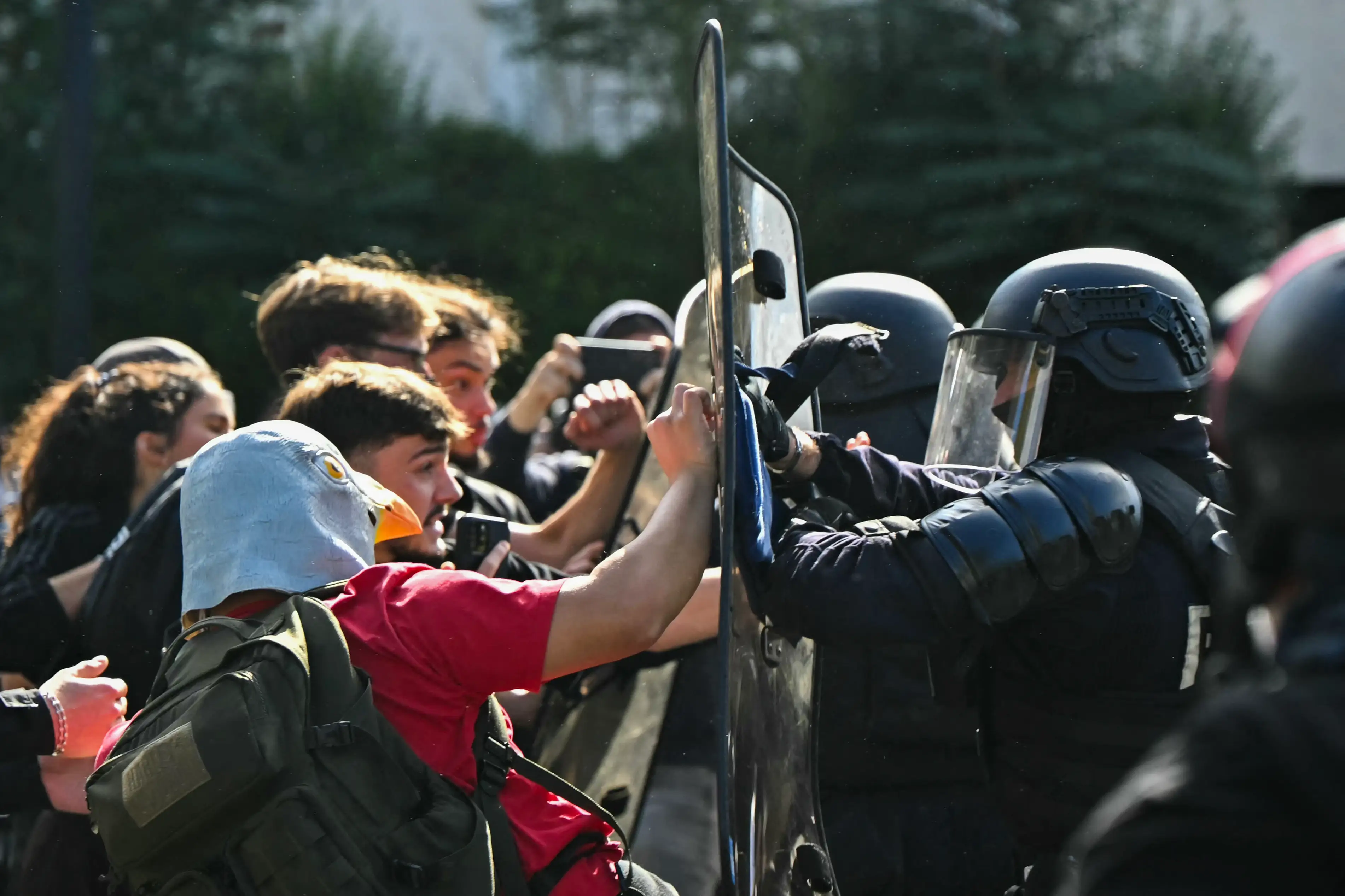 Protesters confront riot police with shields during an outdoor crowd-control scene in bright daylight.