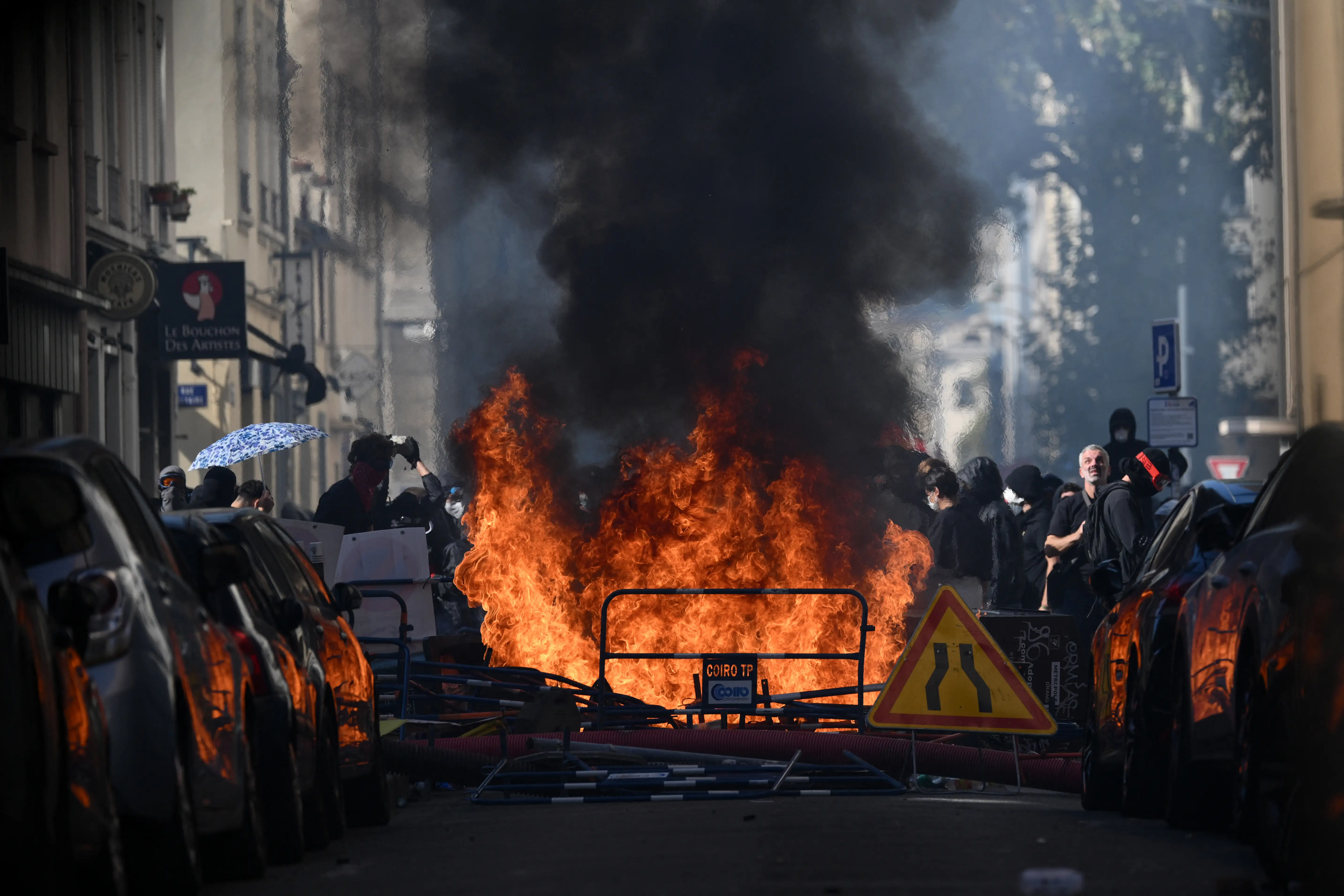 Flames and thick black smoke rise from a street barricade between parked cars as masked people stand nearby.