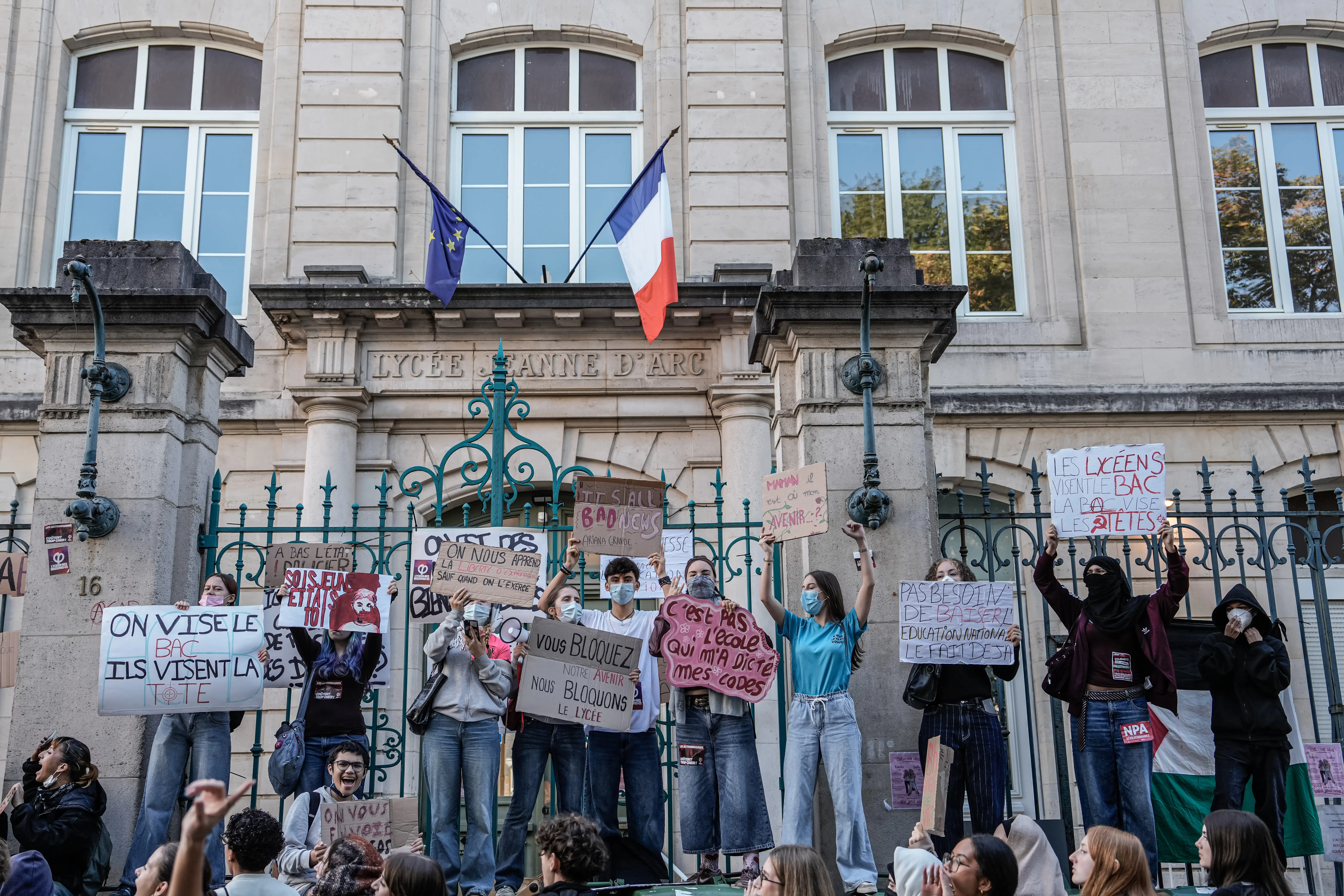 Masked protesters hold French signs outside Lycée Jeanne d'Arc, beneath French and European Union flags.