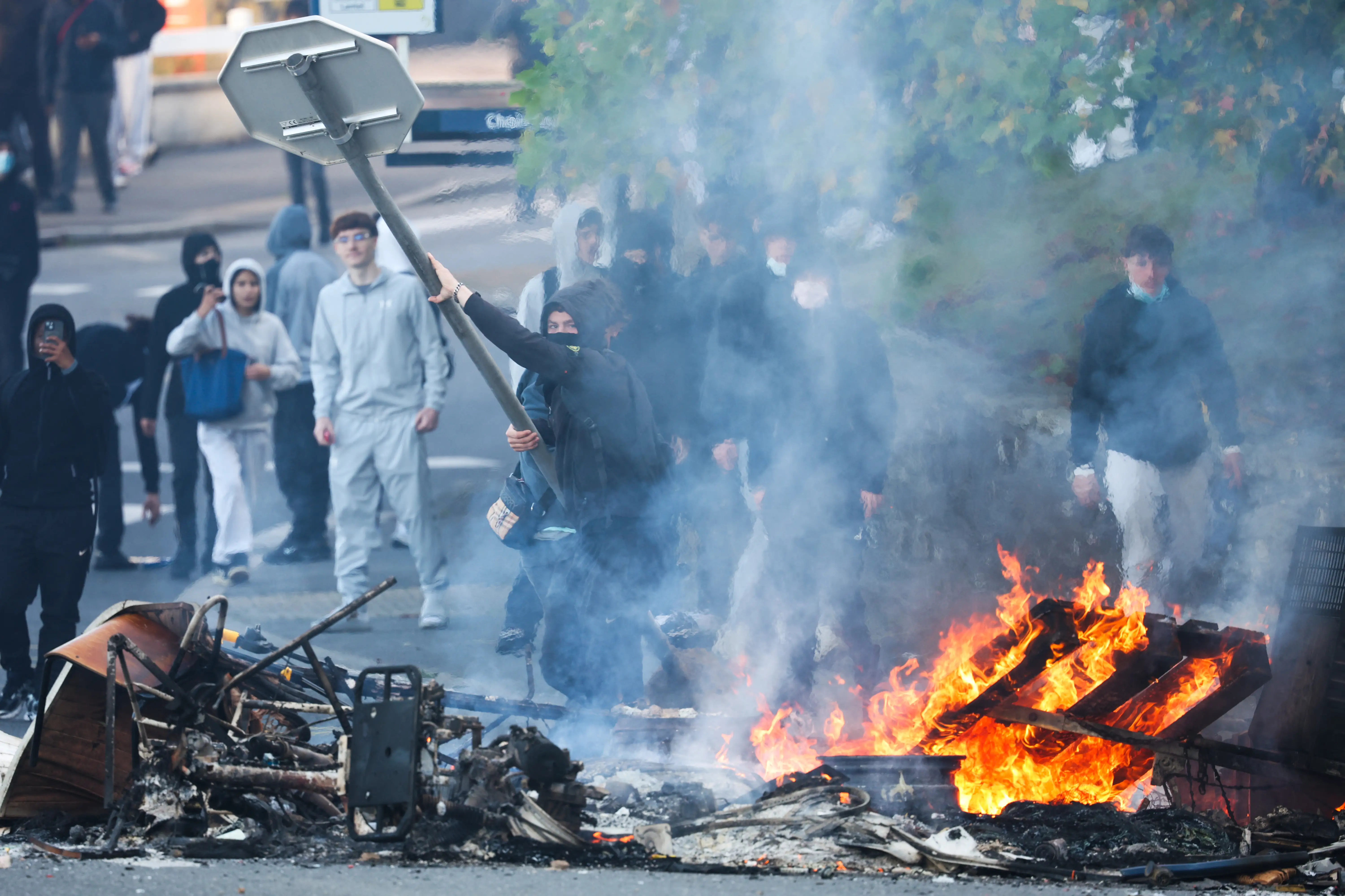 Masked people stand near burning debris and heavy smoke during a street disturbance.