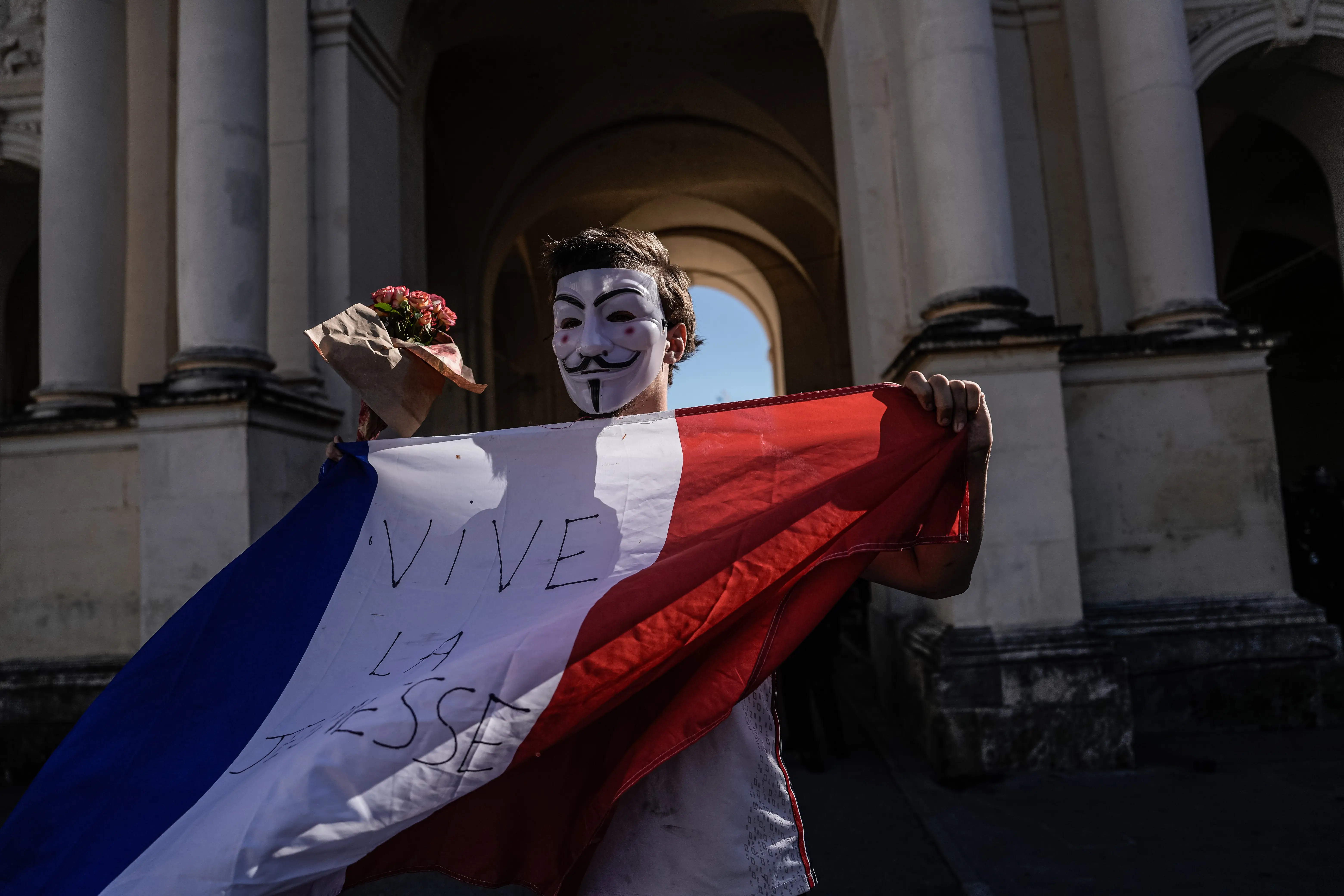 Masked demonstrator holds a French flag with handwritten text in front of a columned stone building.