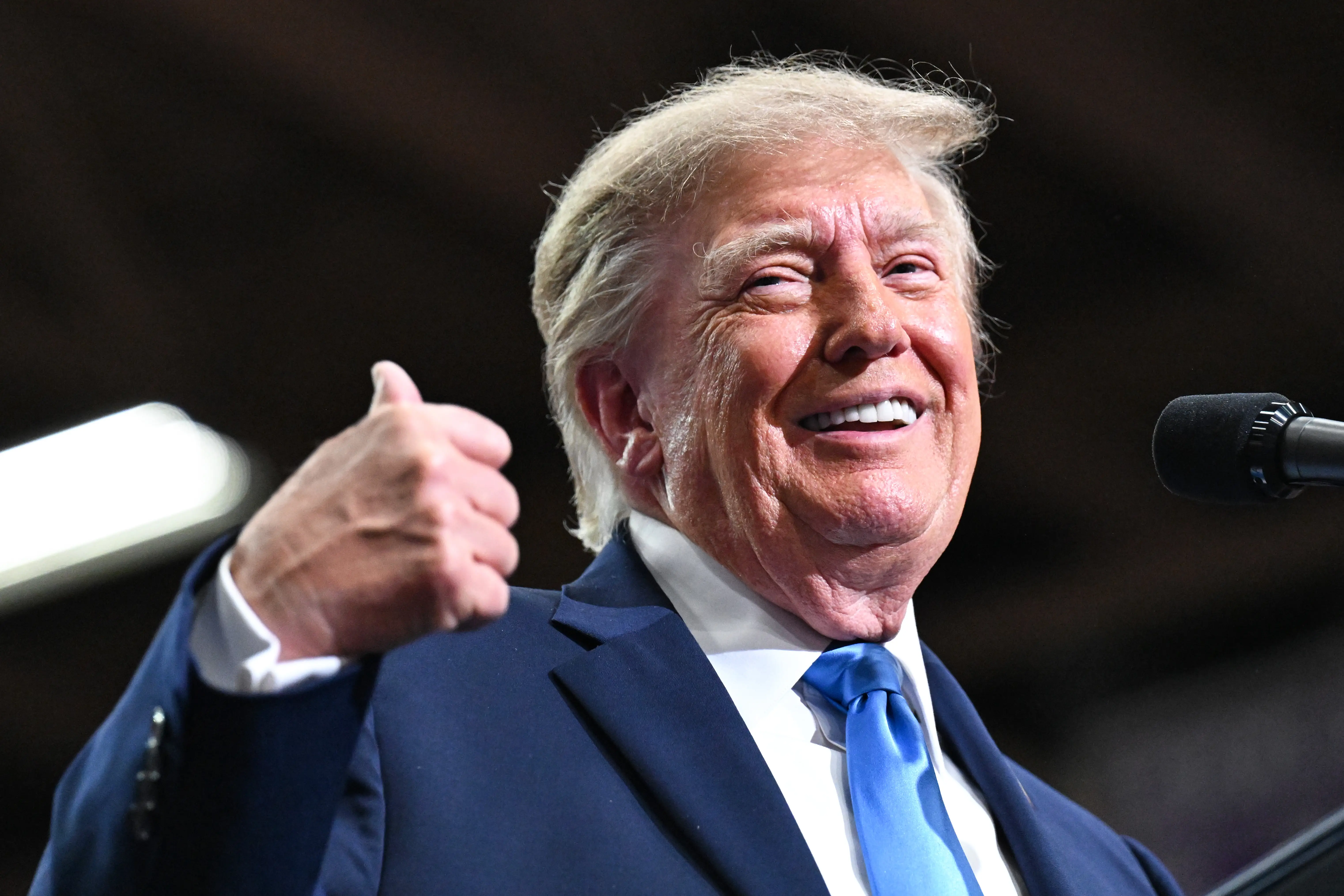 Donald Trump smiles and gives a thumbs-up beside a microphone while wearing a suit and blue tie.