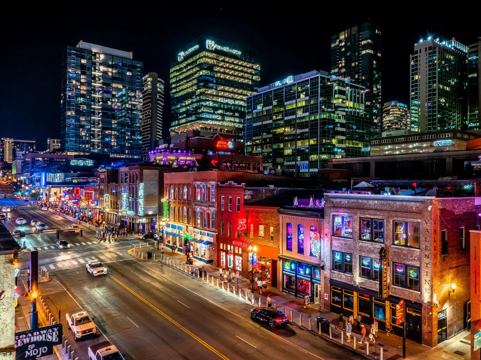 Neon-lit Broadway street in Nashville at night with bars, traffic, pedestrians, and illuminated high-rise buildings.