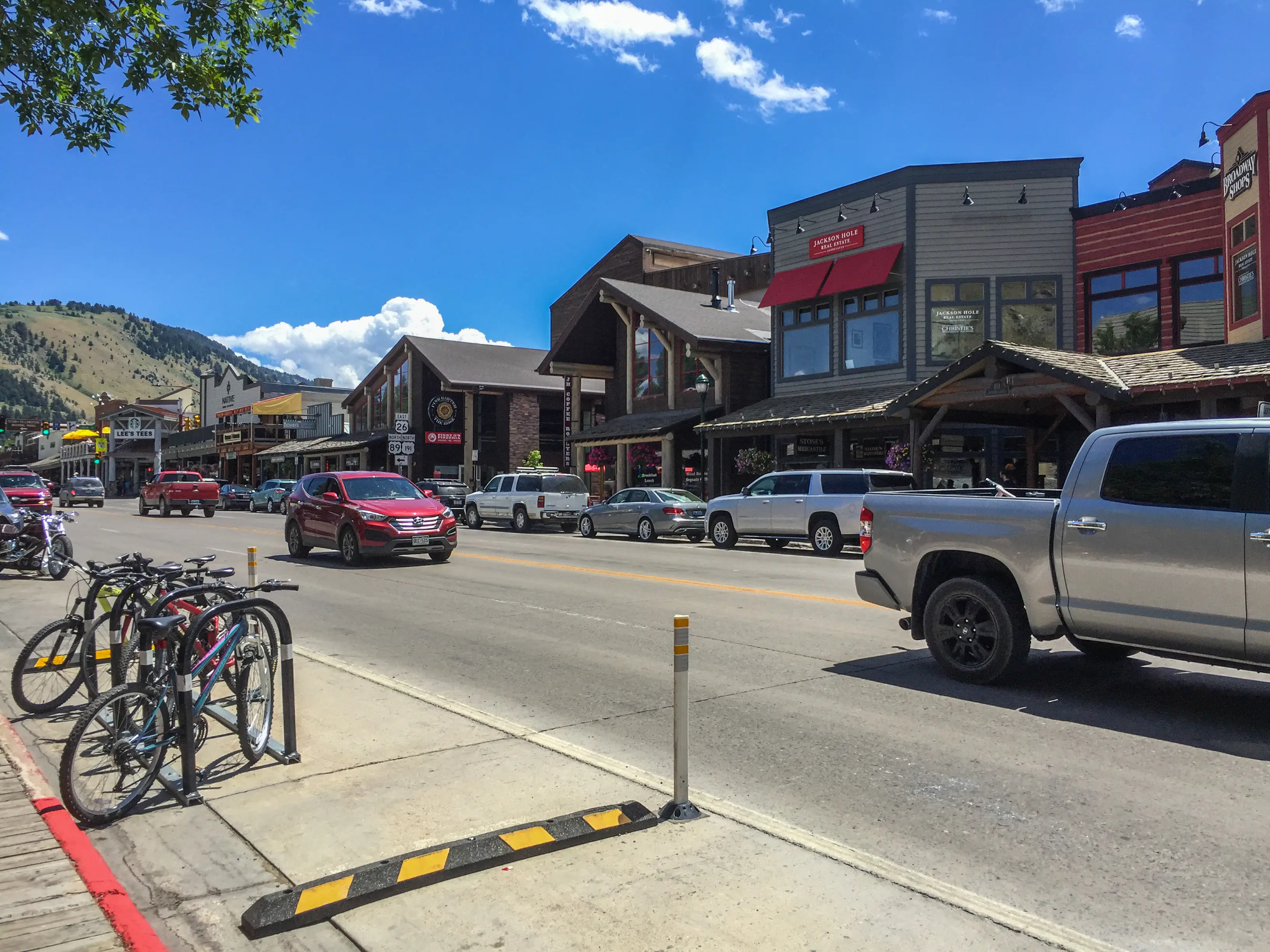 Downtown street with storefronts, parked vehicles, bicycles, and mountains under a bright blue sky.
