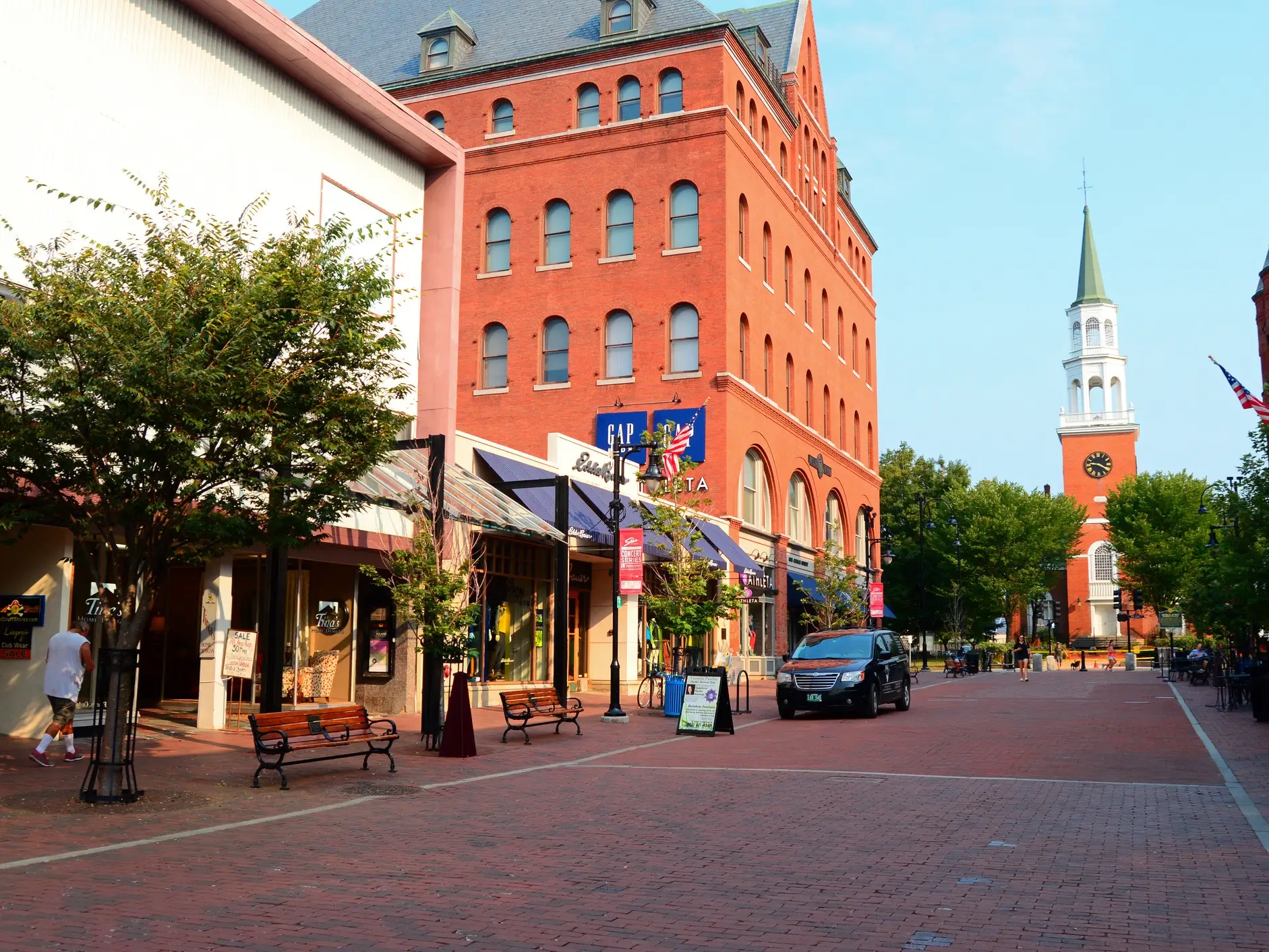 Red-brick pedestrian shopping street with storefronts, benches, a black van, and a church clock tower in the distance.