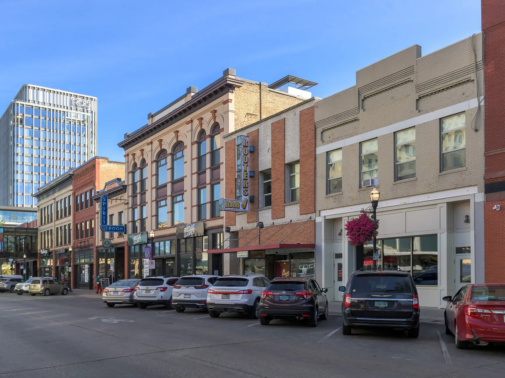 Parked cars line a downtown street with storefronts, bar signs, and a modern high-rise under a clear sky.