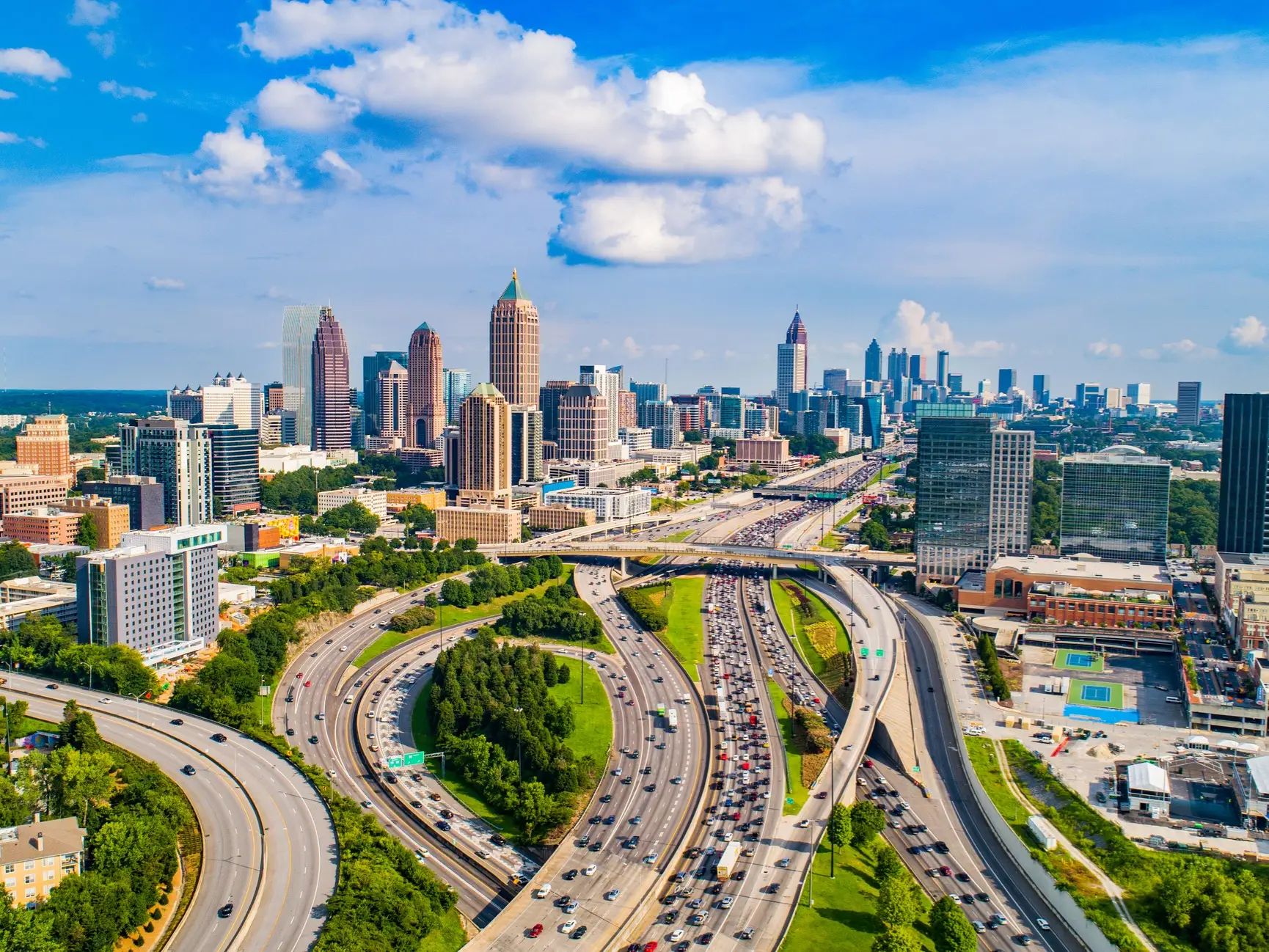Atlanta skyline rises beyond busy multilane highways under a bright blue sky with scattered clouds.