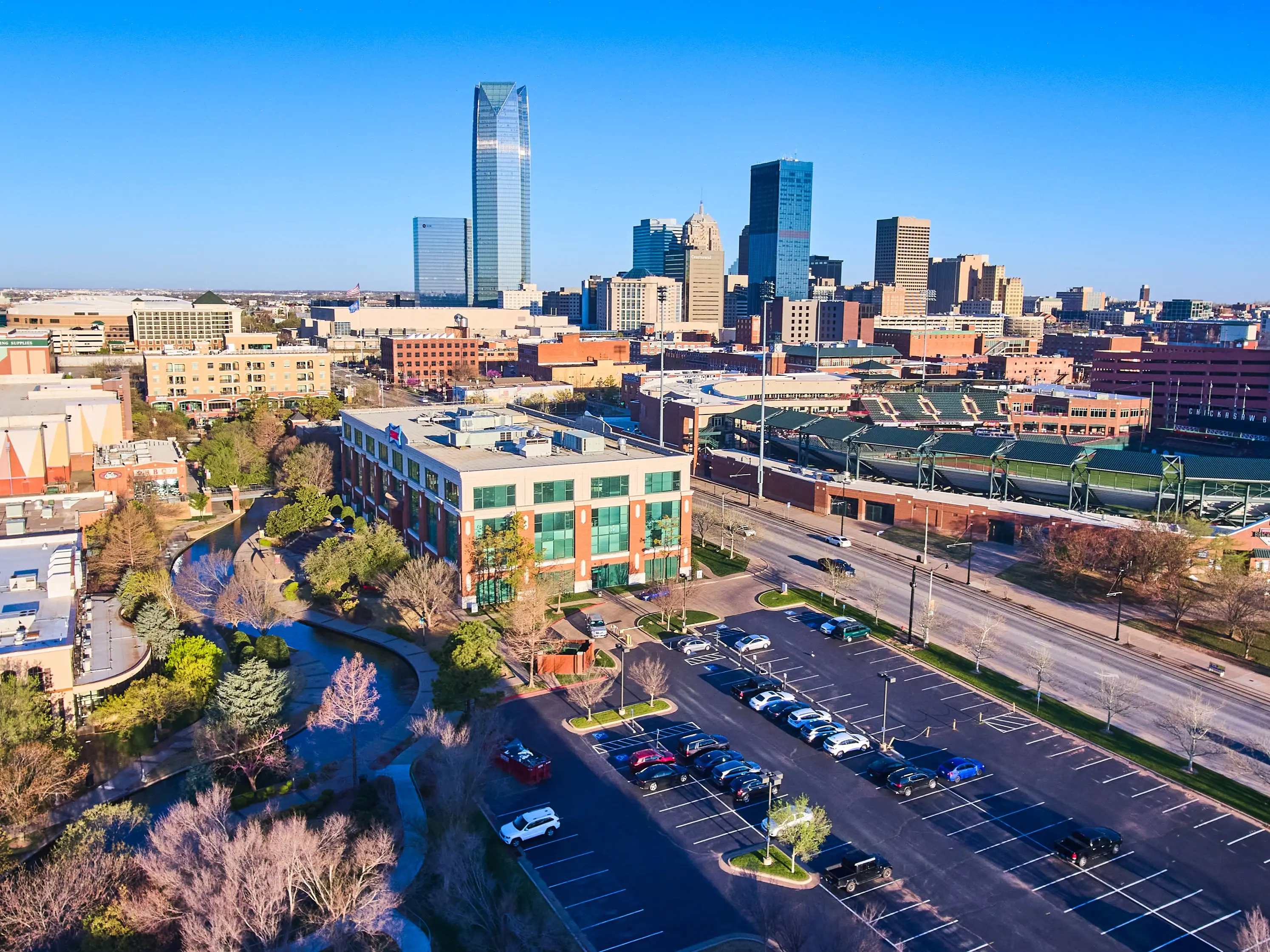 Downtown Oklahoma City skyline with Devon Energy Center, Bricktown ballpark, streets and parking lots under a clear blue sky.