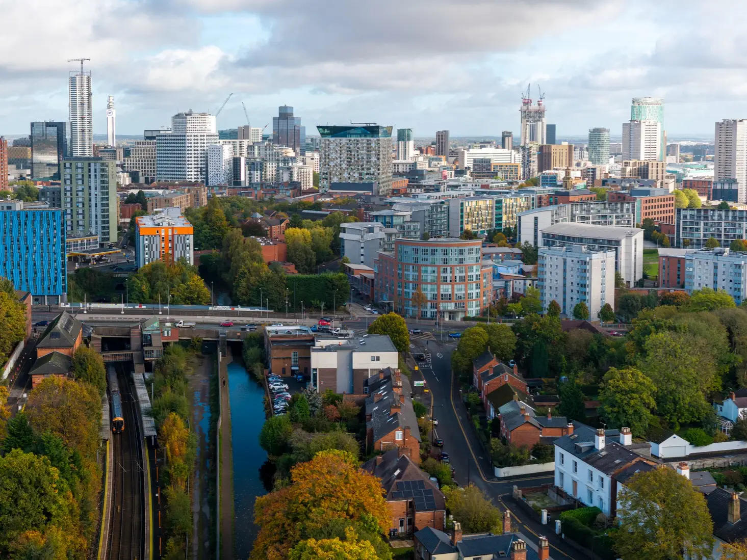 Birmingham city skyline with high-rise buildings, rail tracks, canal, roads, and autumn trees under cloudy skies.