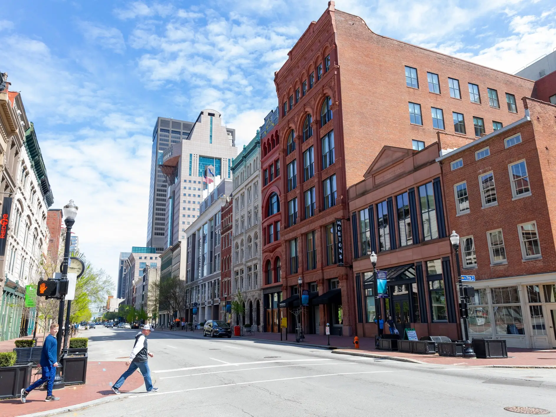 Historic brick buildings line a downtown street with pedestrians crossing under a partly cloudy blue sky.