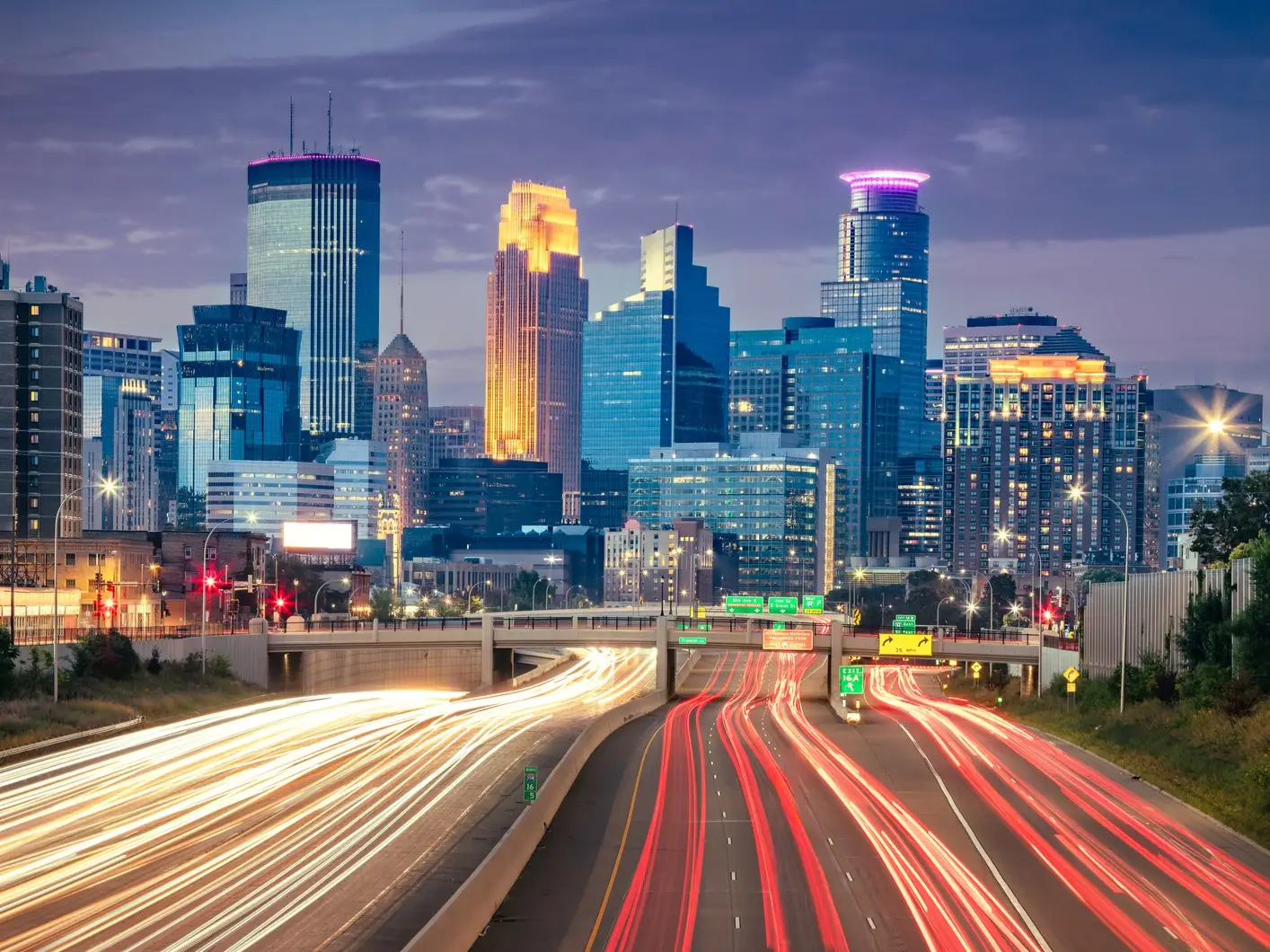 Downtown Minneapolis skyline glows at dusk above freeway lanes with long-exposure red and white traffic trails.