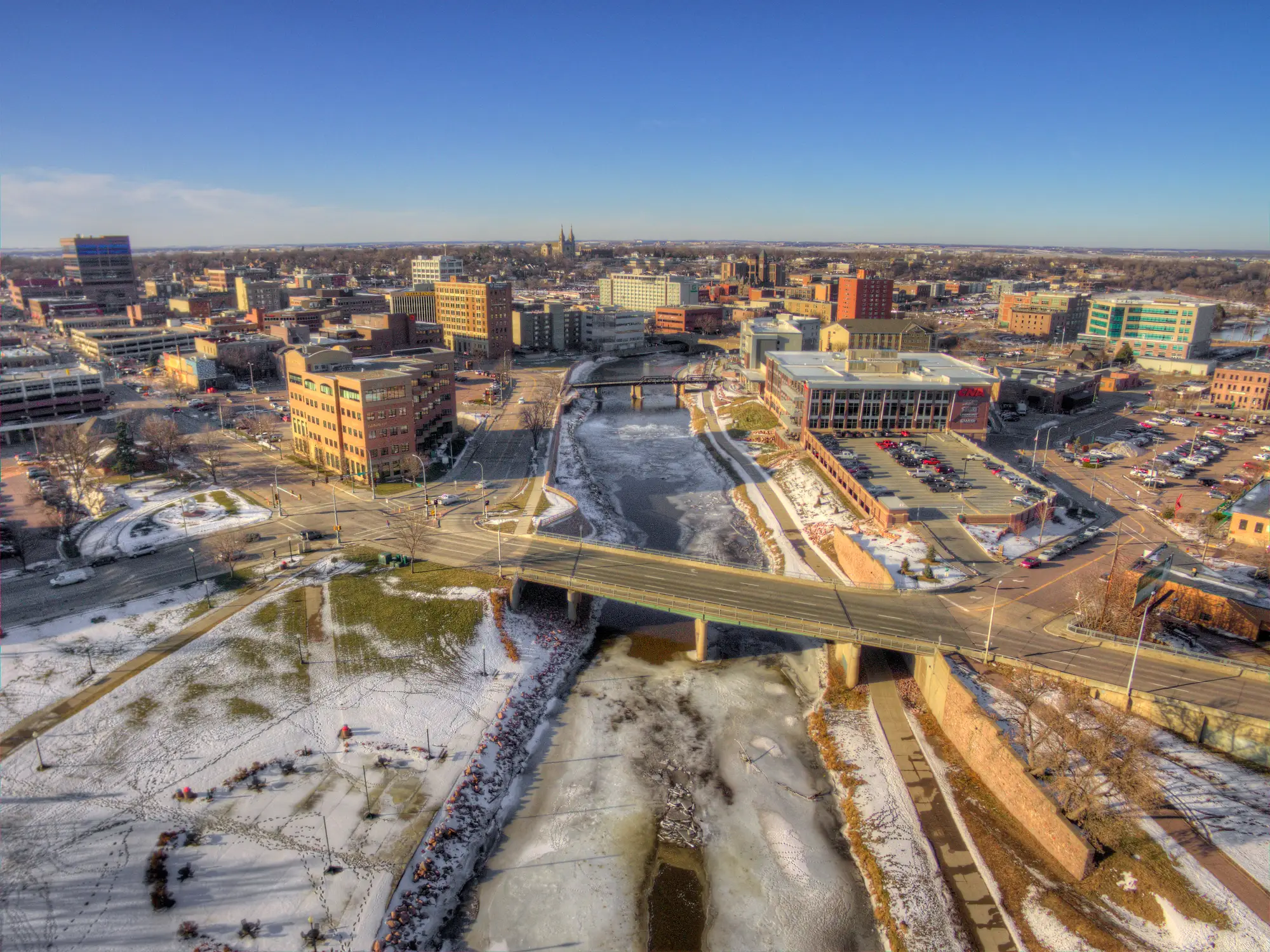Snow-dusted downtown South Bend skyline surrounds the partially frozen St. Joseph River beneath a clear blue sky.