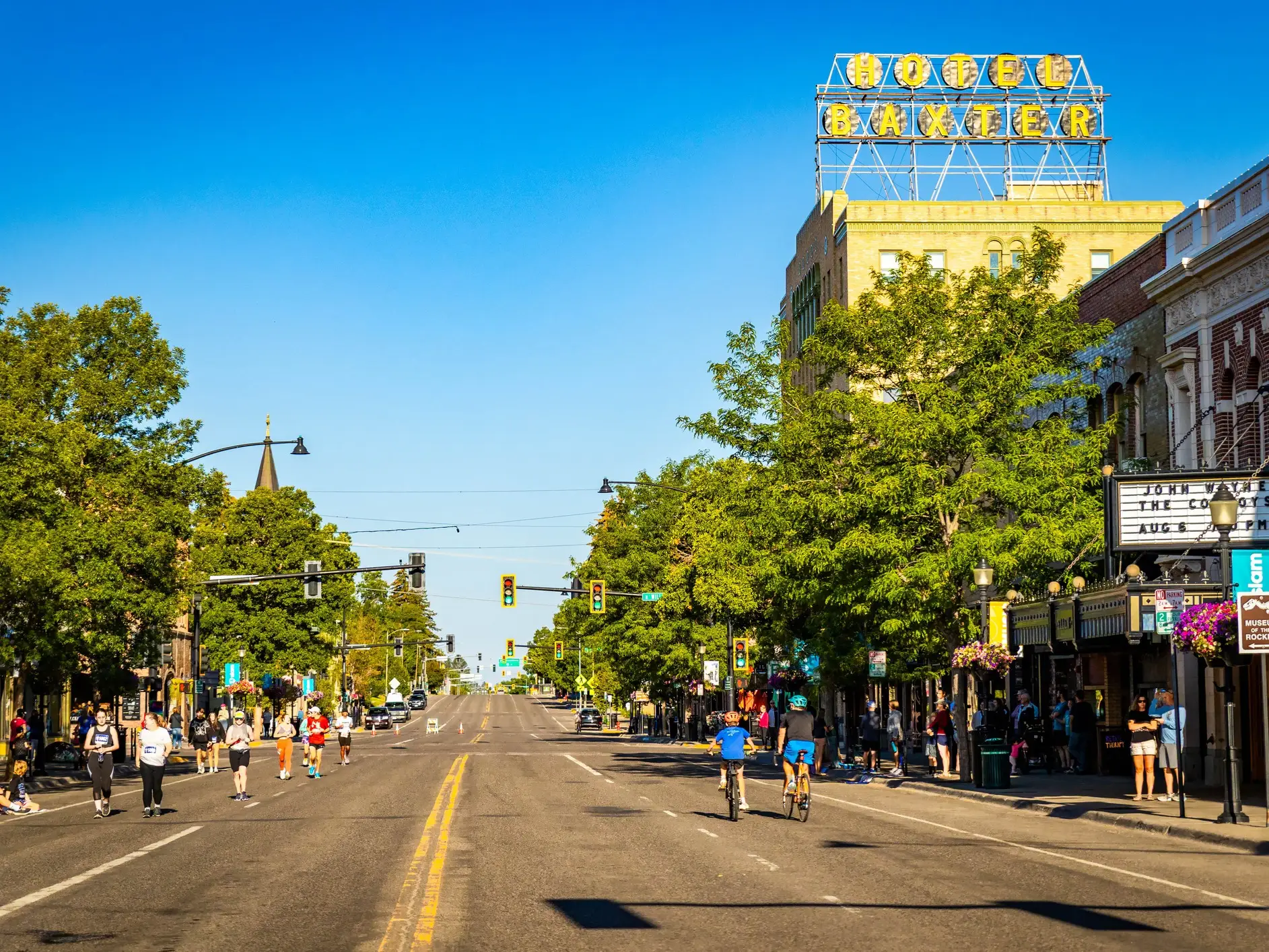 Runners and cyclists travel along a sunny downtown street beneath the Hotel Baxter rooftop sign.