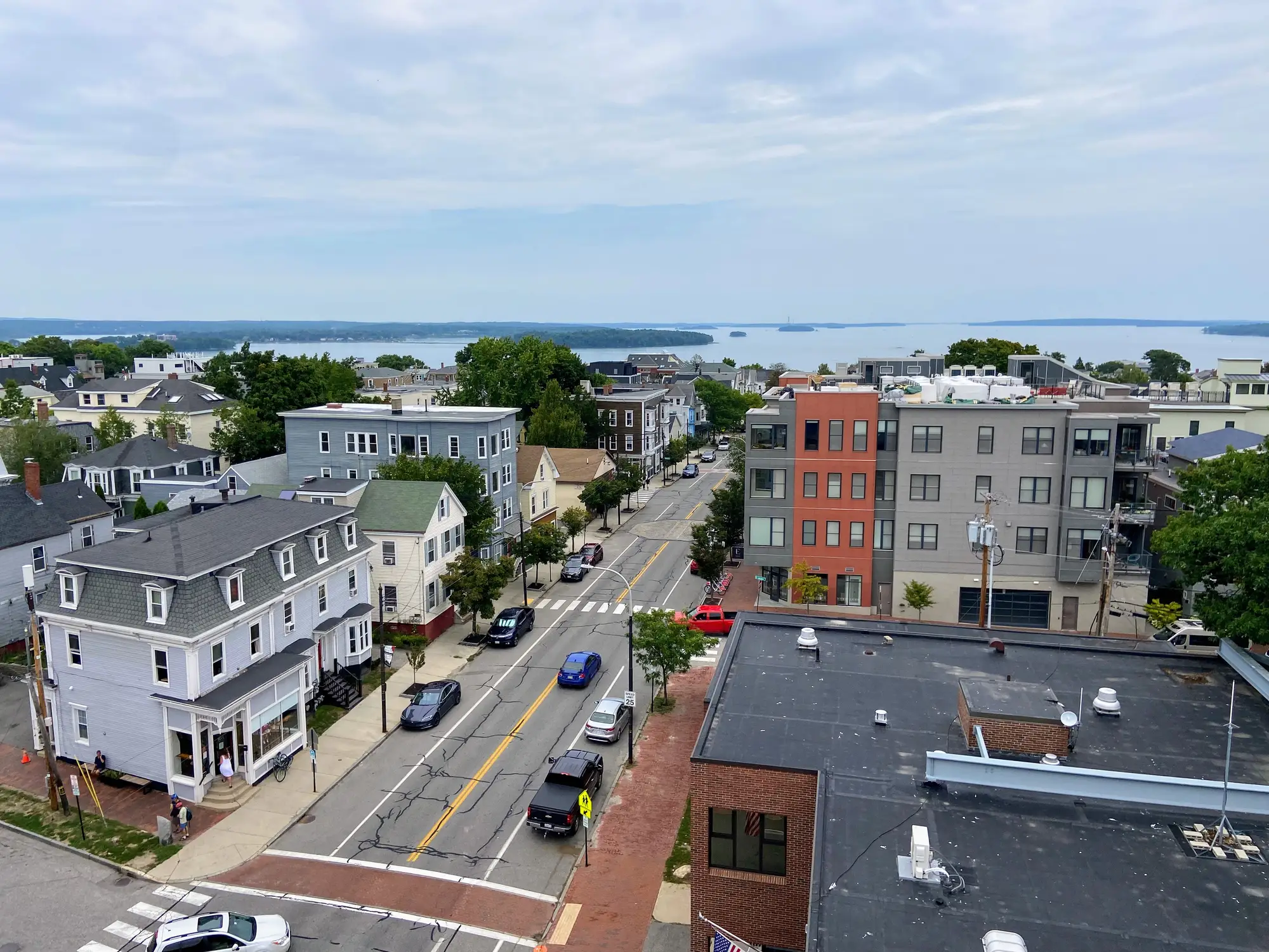 Elevated view of a coastal neighborhood street with houses, apartment buildings, parked cars, and a bay in the distance.