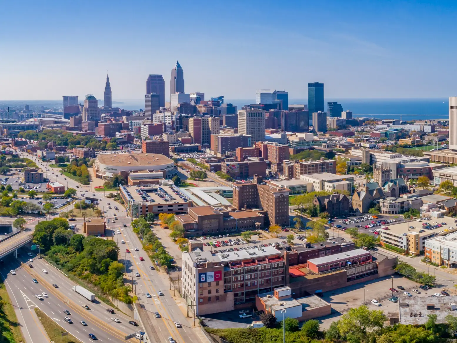 Aerial view of downtown Cleveland skyline with high-rises, roads, parking lots, and Lake Erie in the background.