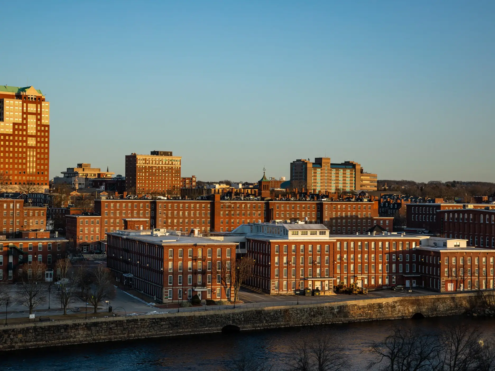 Red brick waterfront buildings and a city skyline are lit by warm sunlight beneath a clear blue sky.