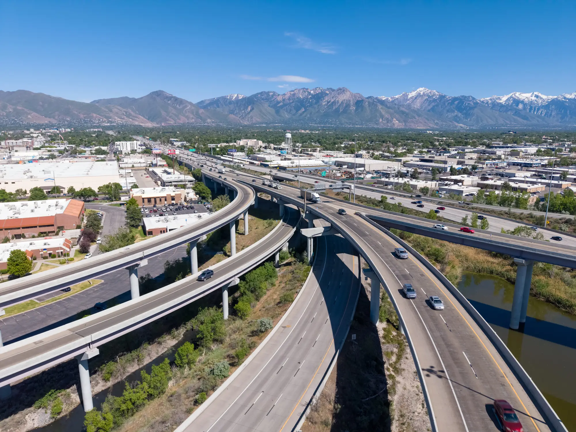 Elevated freeway ramps curve through an urban industrial area with vehicles, waterways, and mountains in the distance.