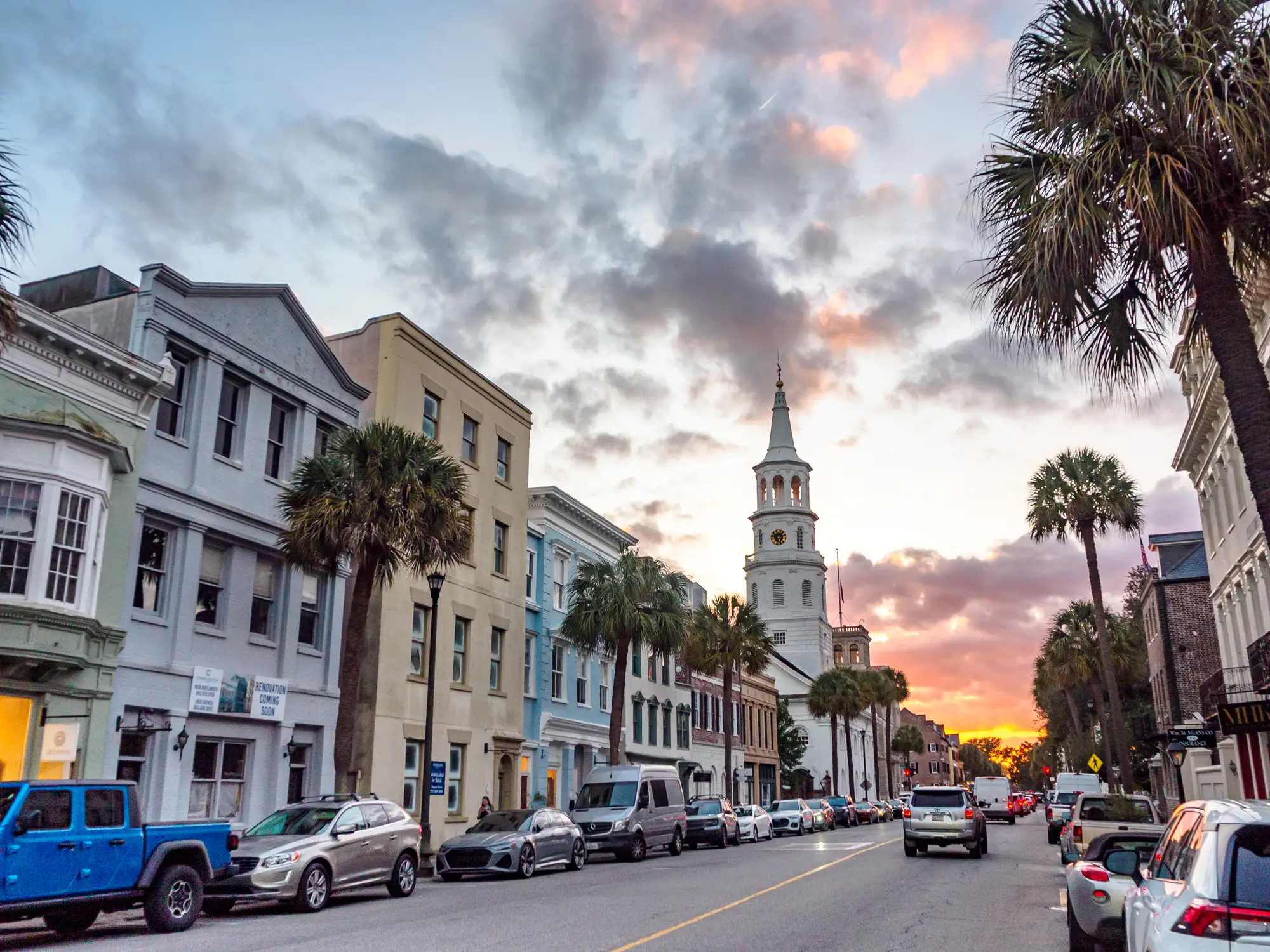 Historic downtown street with palm trees, parked cars, and a white church steeple beneath a colorful sunset sky.