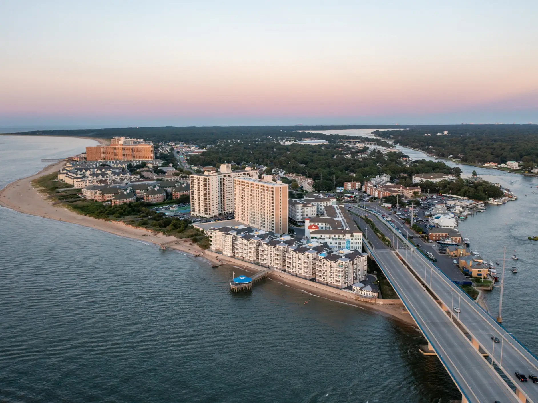 Aerial view of beachfront high-rise buildings, a bridge, and waterways along a coastal neighborhood at sunset.